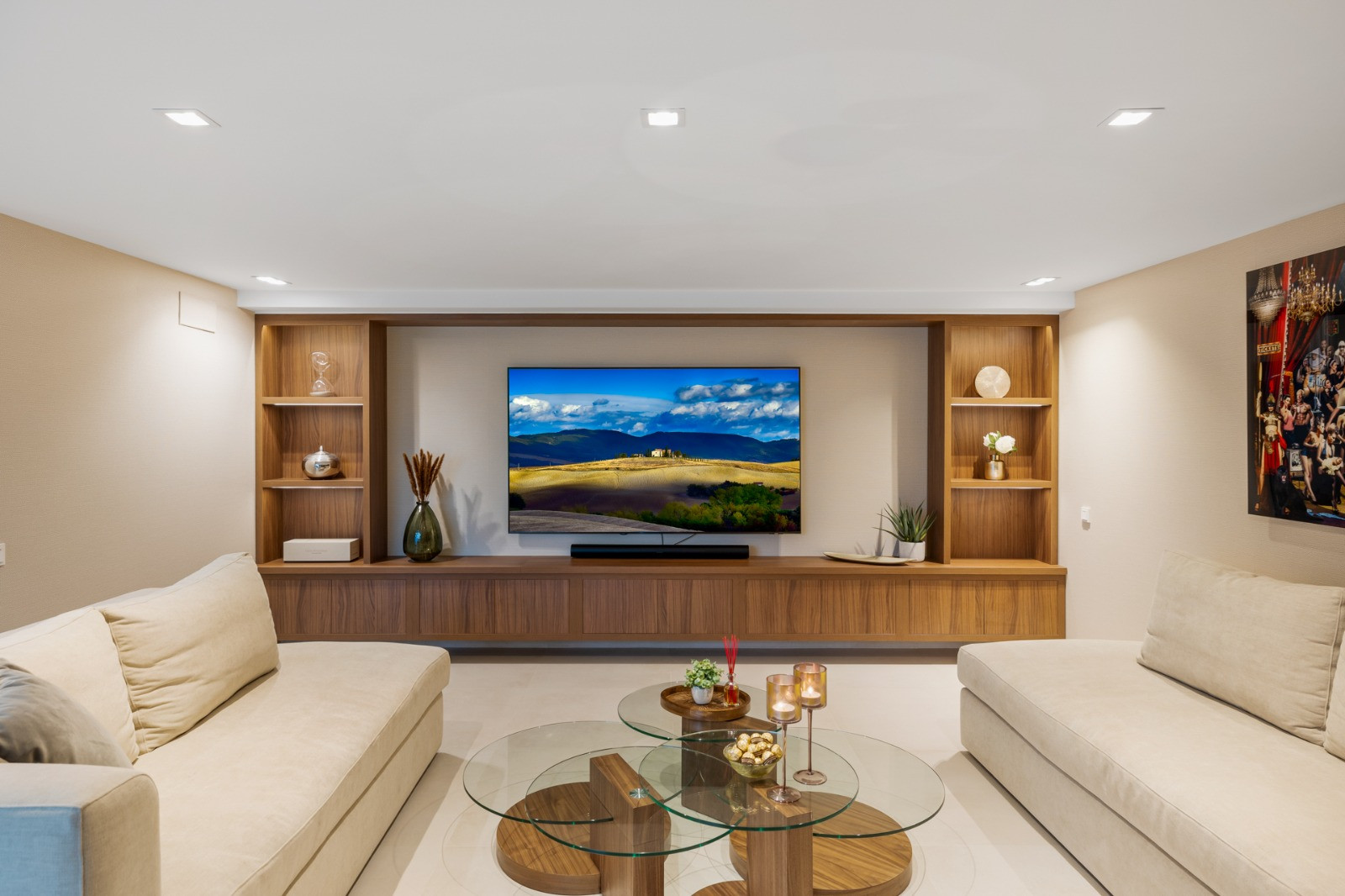 Living room with light-colored sofas and a wall-mounted TV. Wooden shelves and decorative items visible.