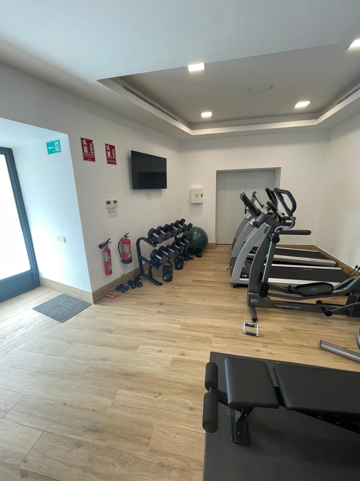 Bright gym area with dumbbells and cardio machines on wooden flooring.