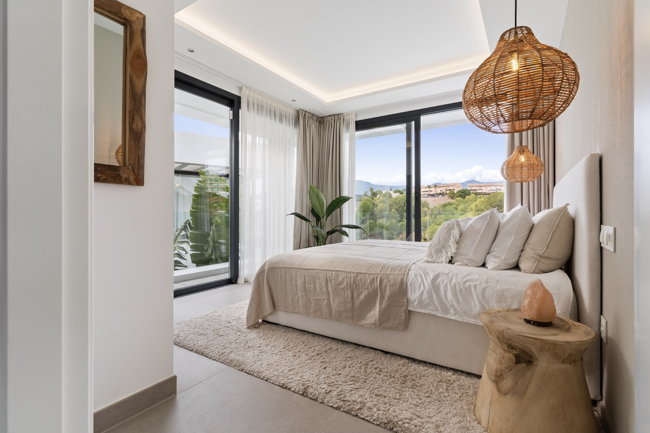 Bright bedroom with large windows, bedding, and decorative plants.
