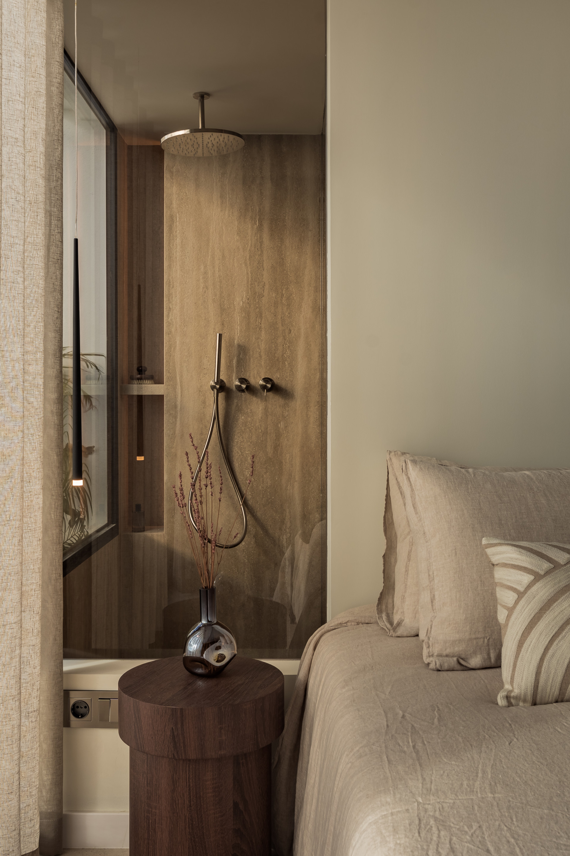 Bedroom corner with a bed, dark wood nightstand, and an open concept shower visible through glass.