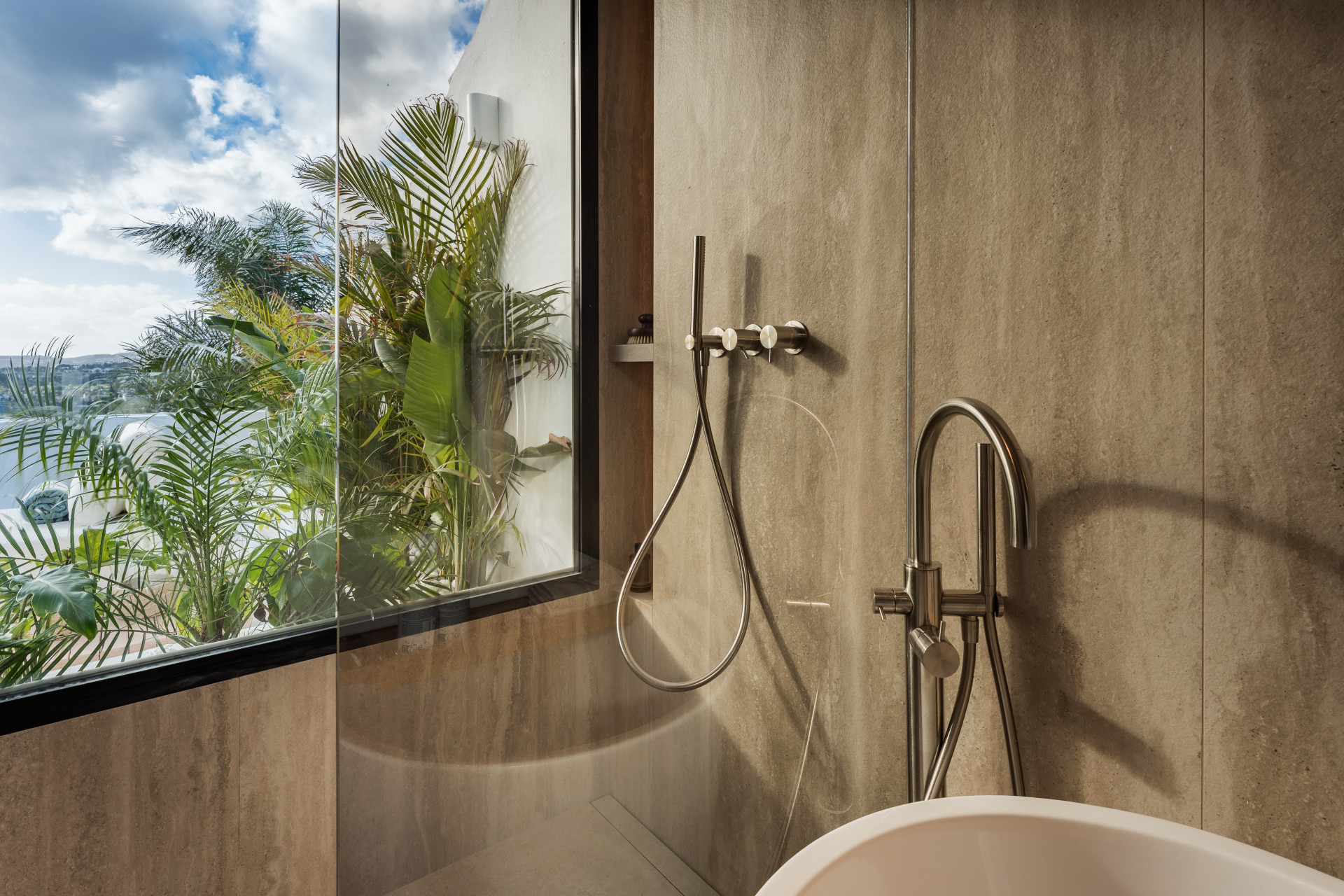 Bathroom shower area with large window overlooking lush tropical foliage and blue skies.