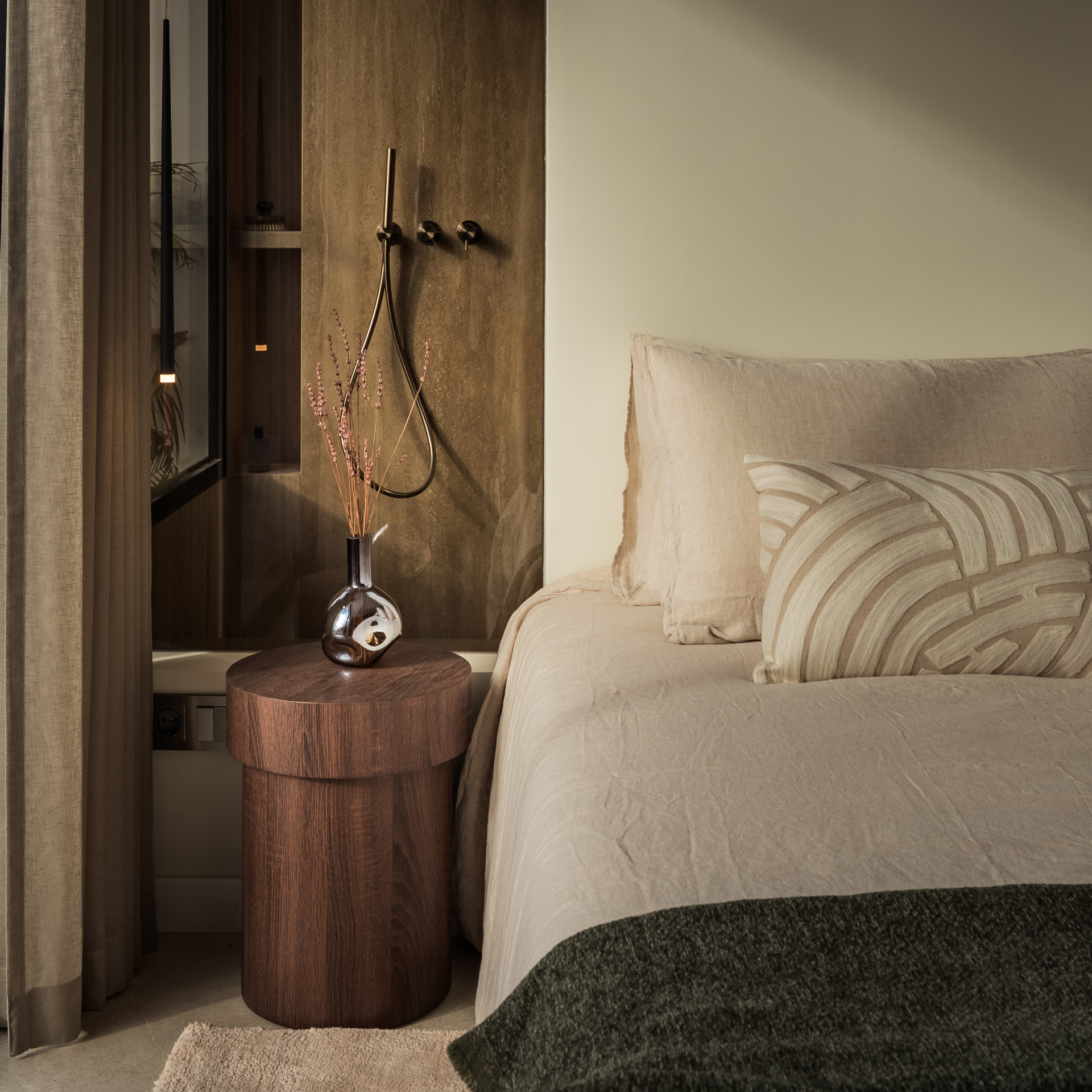 Bedroom detail showing a bed with linen bedding, a dark wood nightstand, and a vase with dried flowers.