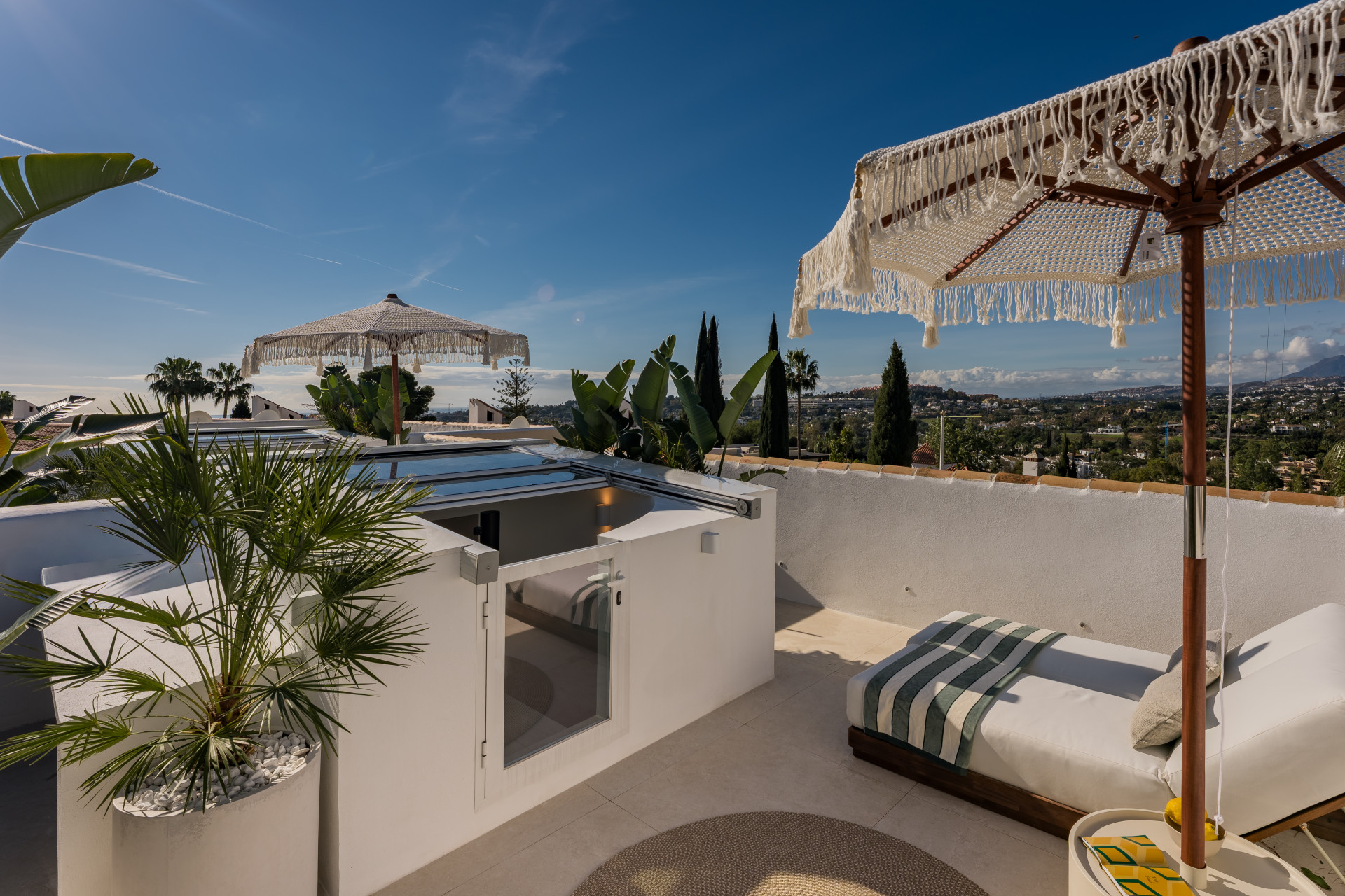 Rooftop terrace with two lounge chairs, decorative parasols, potted plants, and a partial view of a skylight.