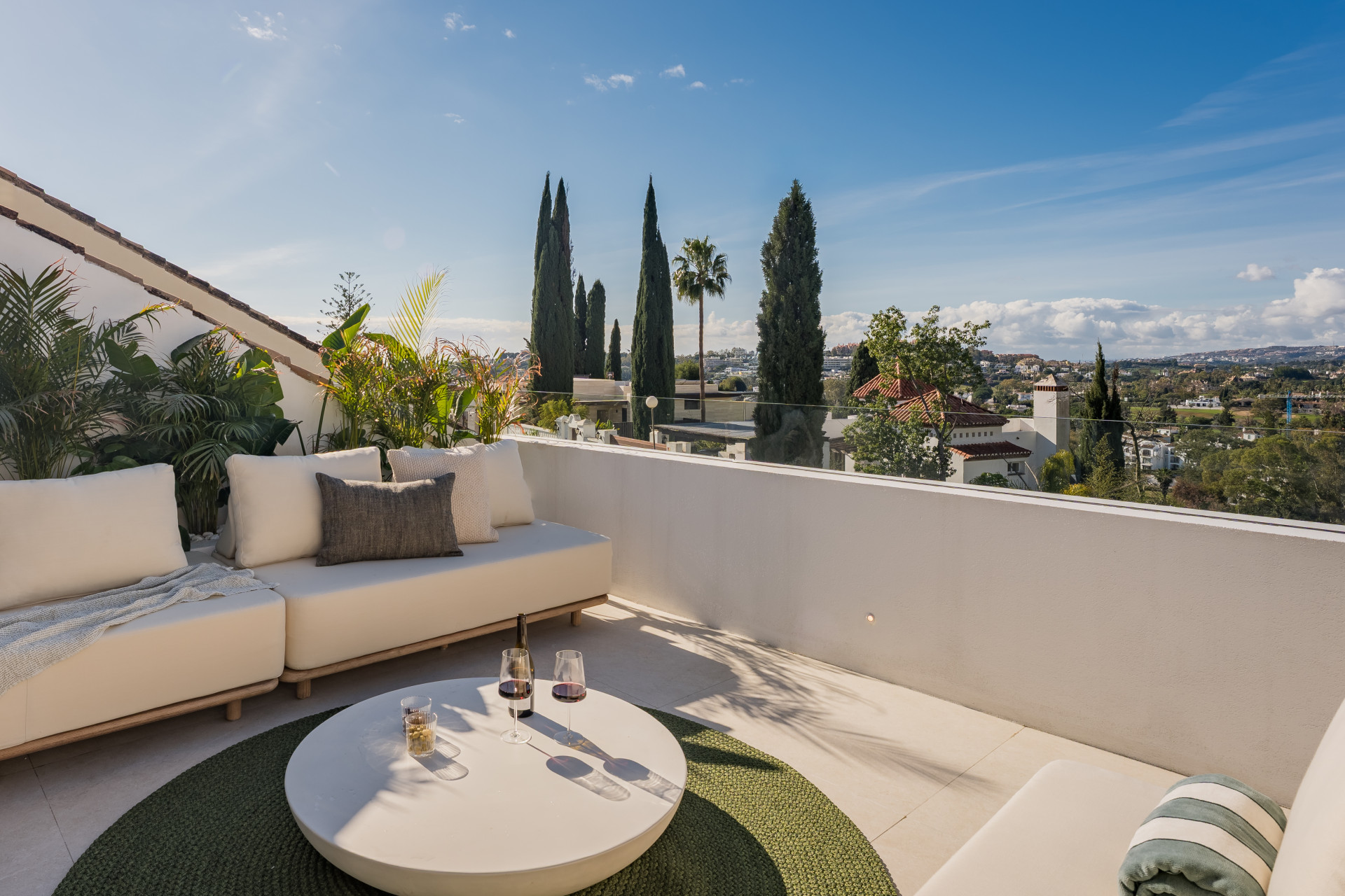 Outdoor terrace featuring a white sectional sofa, a round coffee table, and distant city views under a blue sky.
