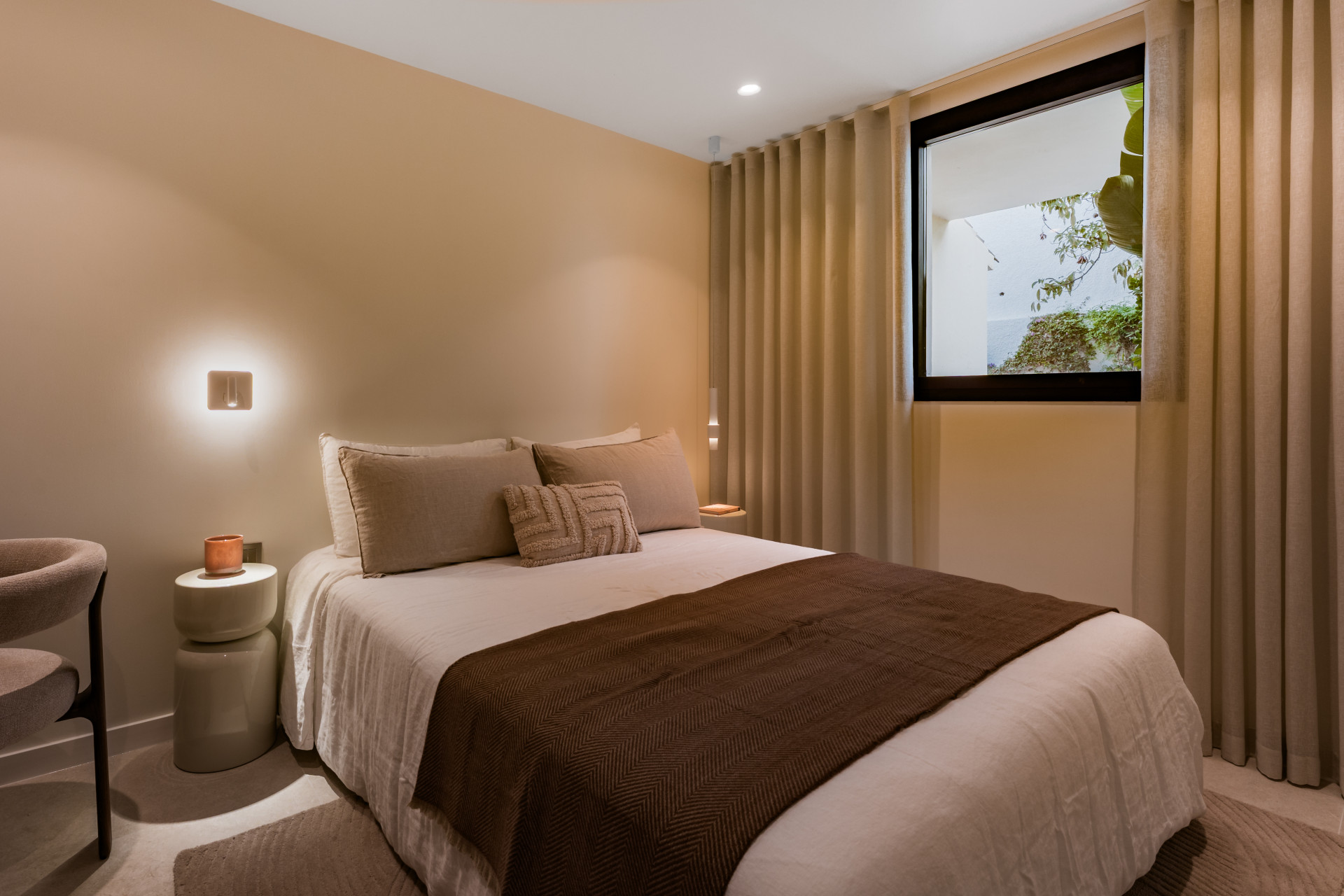 Bedroom with a large bed, two nightstands, a wall lamp, and a window offering a view of greenery.