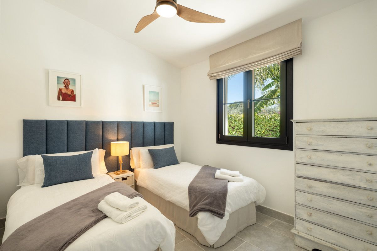 Bedroom with two single beds, blue headboards, a ceiling fan, and a window overlooking greenery.
