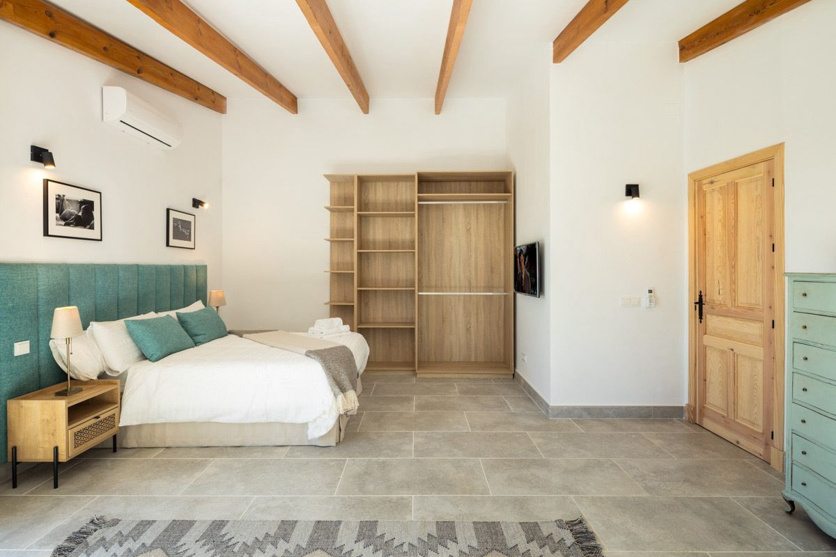 Bedroom with teal headboard, white bed, open wardrobe, light gray tiled floor, and wooden door.