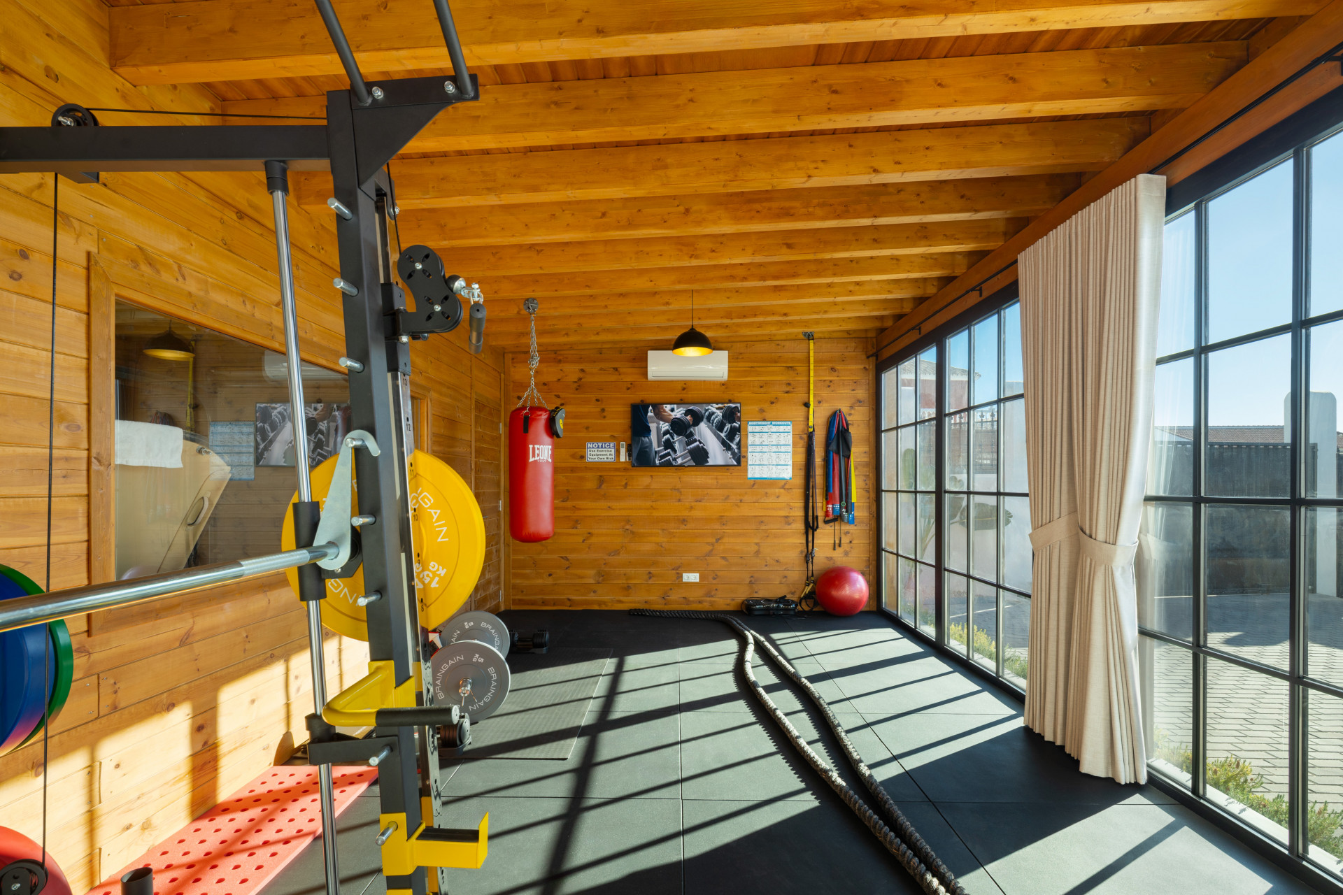 Gym room with wooden walls and ceiling, a weight rack, punching bag, and large windows with black frames.