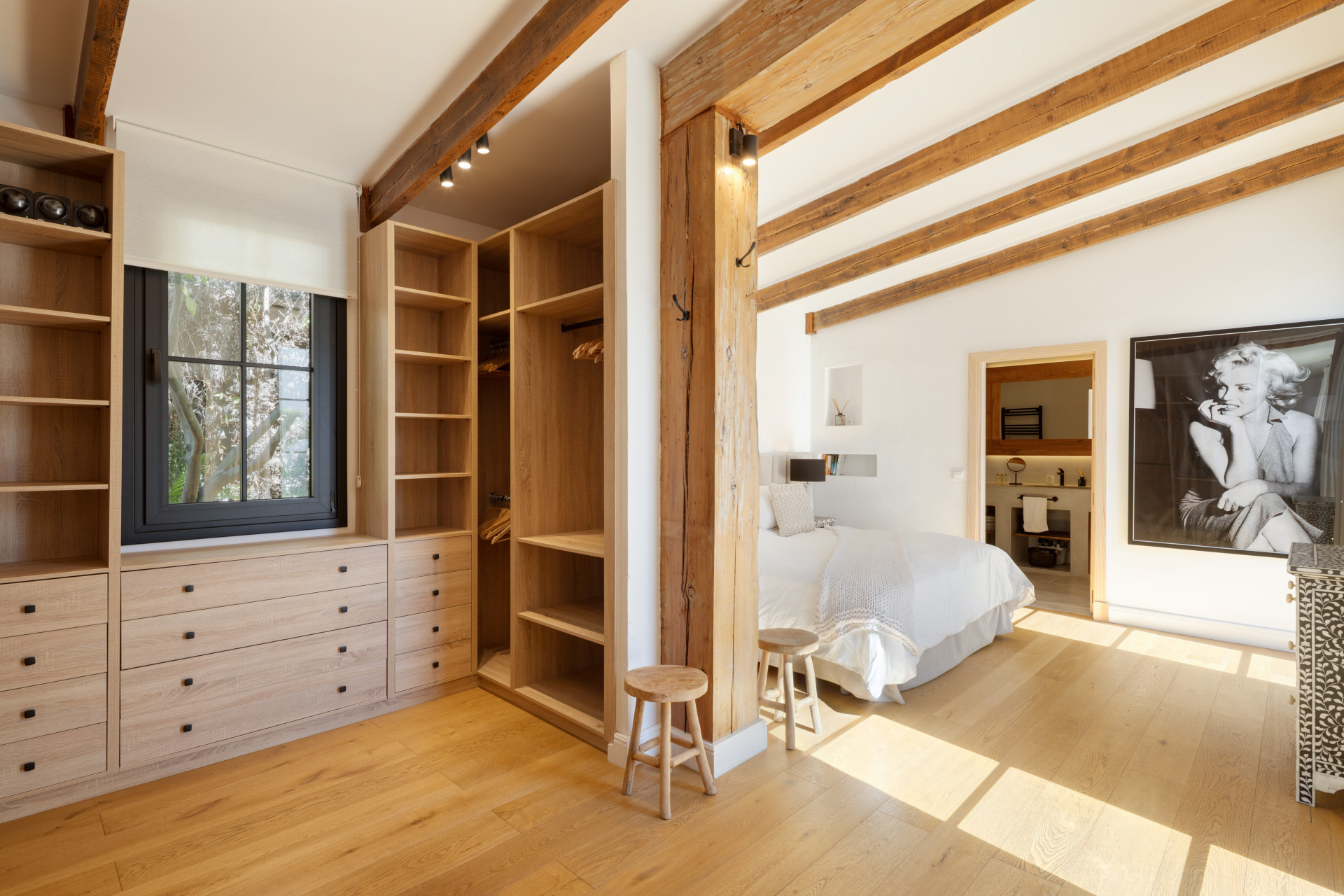 Bedroom with walk-in closet, wood floors, exposed beams, and a view into a modern bathroom.