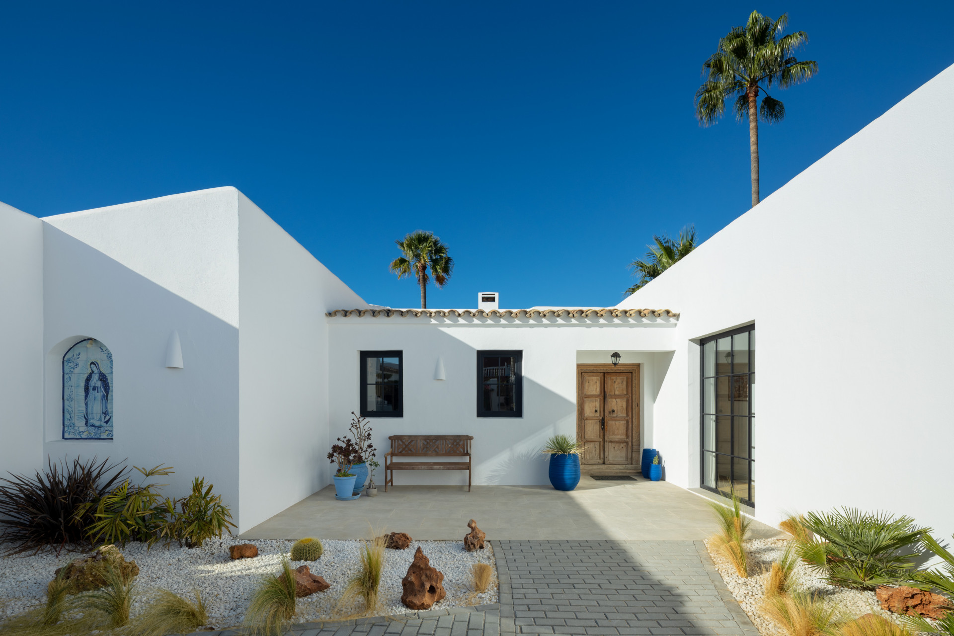 Outdoor courtyard with white walls, a tiled Virgin Mary plaque, a wooden bench, and a carved double door.