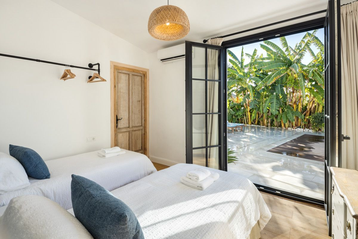Bedroom with two single beds, an open glass door leading to a patio with banana trees, and a wooden door.