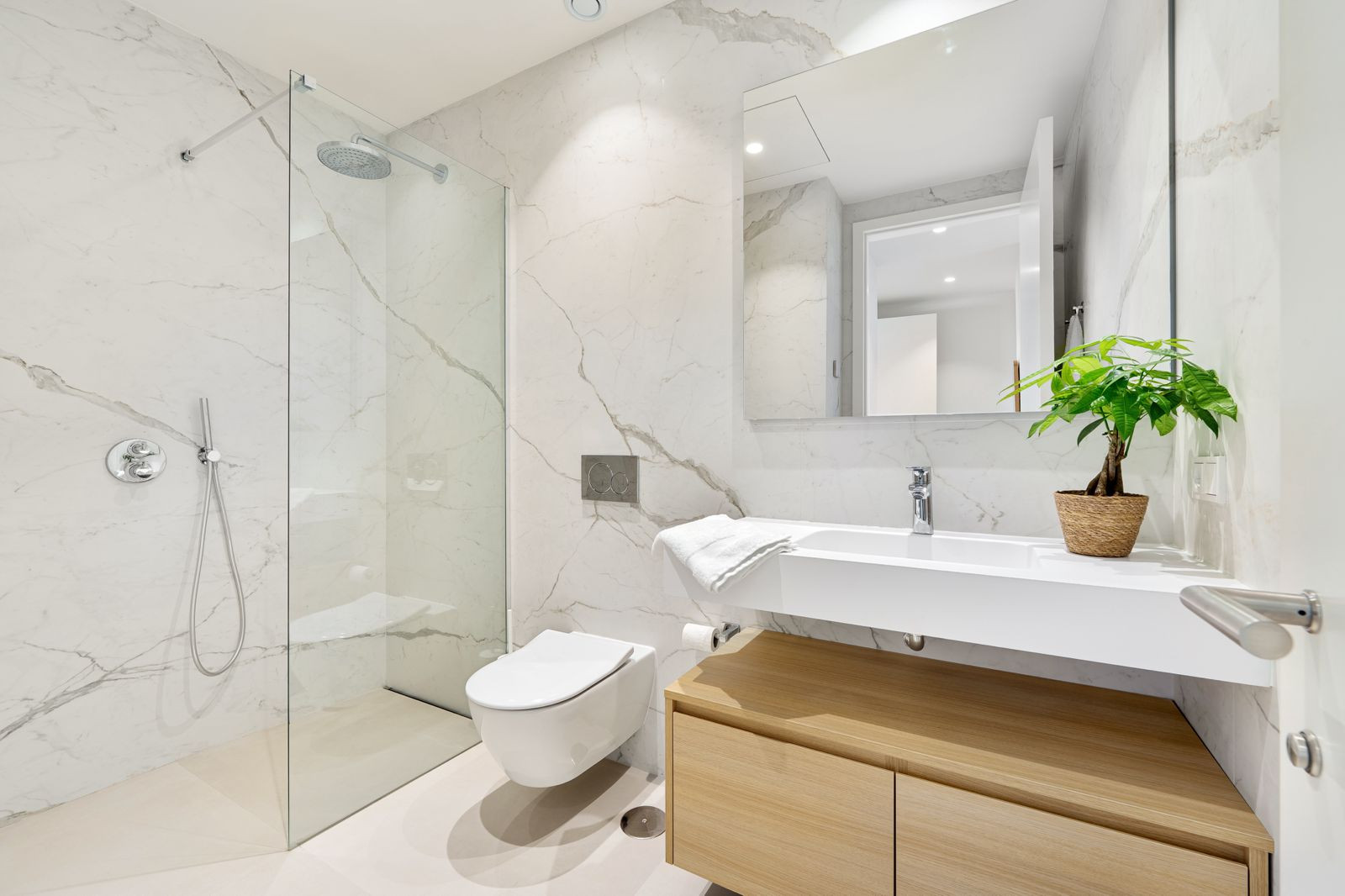 Bathroom with marble walls, a glass-enclosed shower, wall-mounted toilet, and wood vanity with sink.