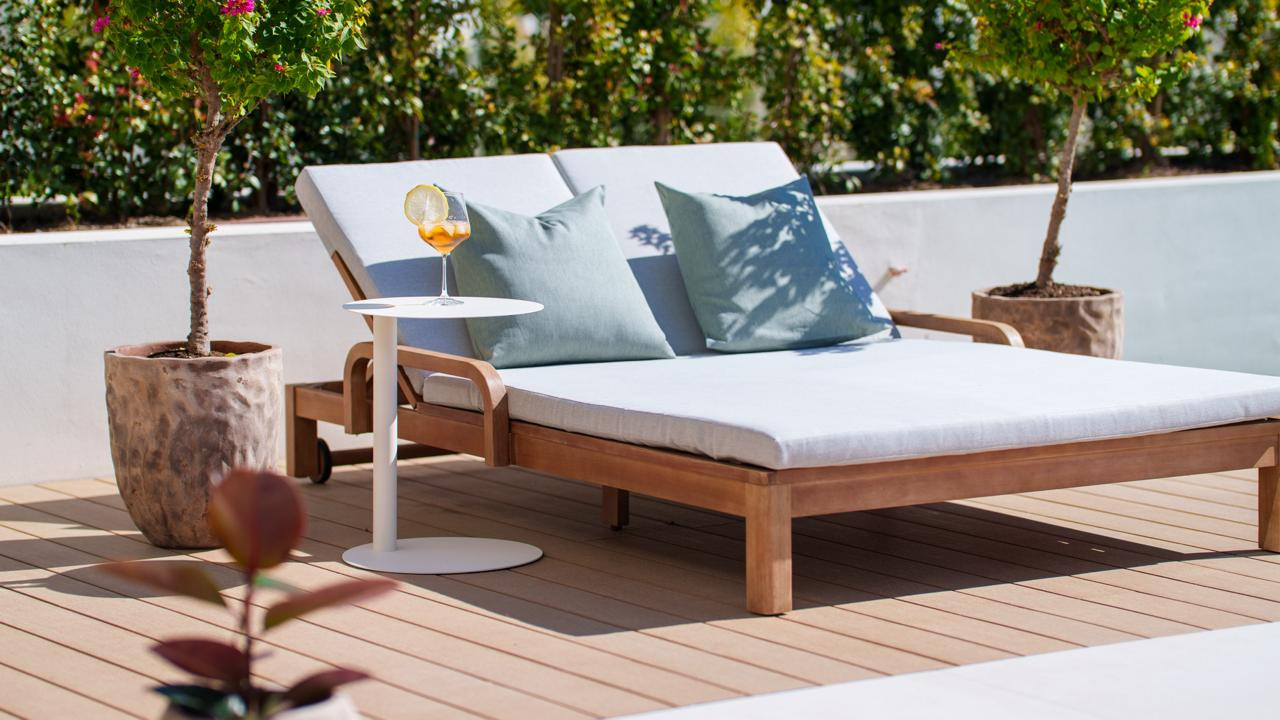 Close-up of a wooden double lounge chair with cushions and a drink on a side table on a deck.