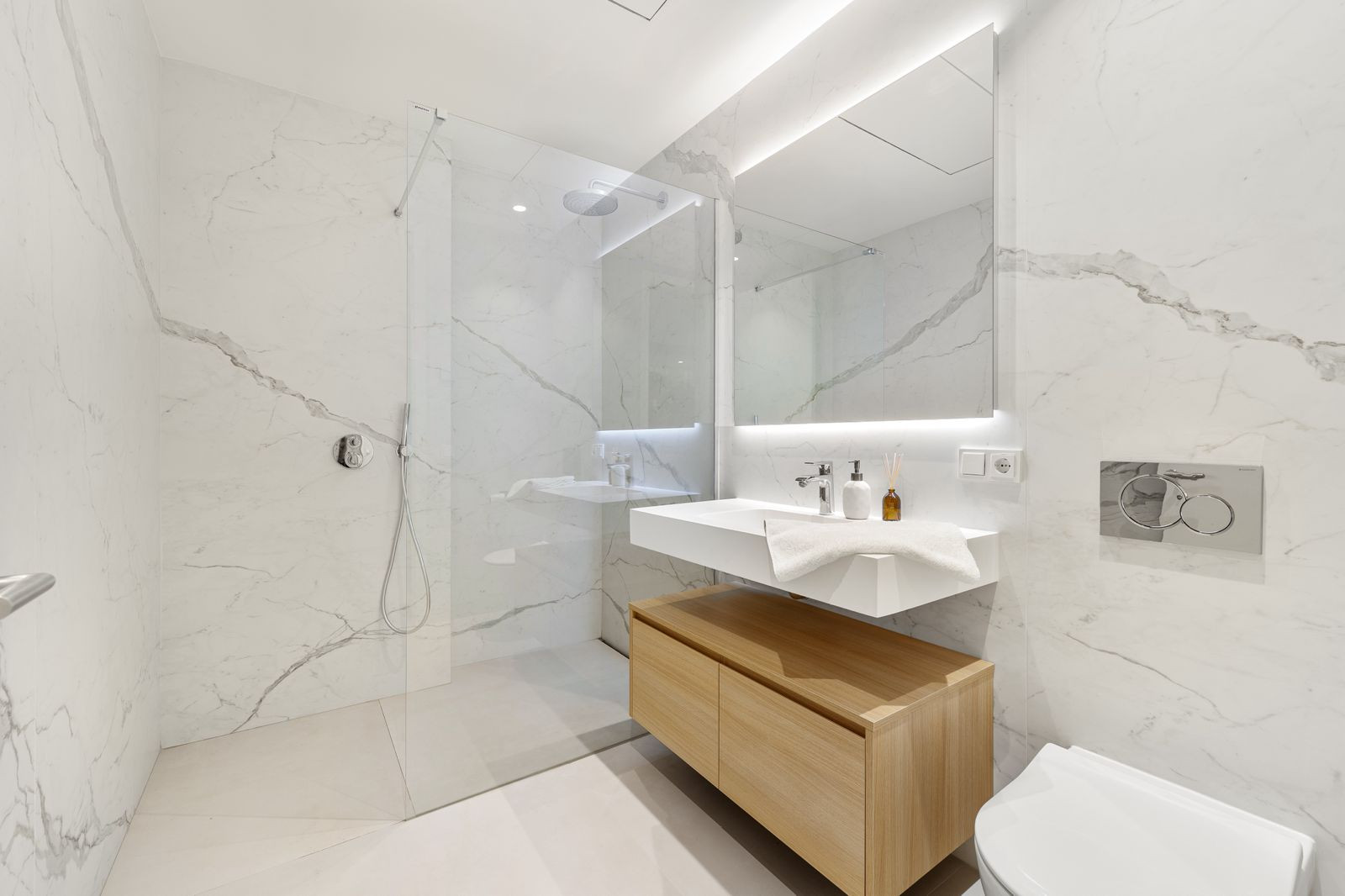 Modern bathroom with white marble walls, walk-in glass shower, floating vanity, and mirror with backlighting.
