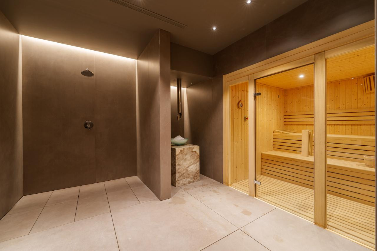 Spa area with a wooden sauna cabin, a rain shower, and a stone pedestal holding a bowl, all in warm tones.