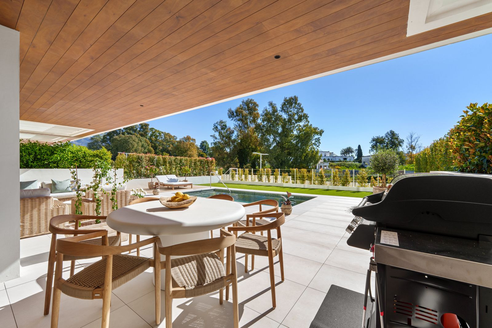 Spacious covered patio with a dining table, grill, and swimming pool visible under a clear blue sky.