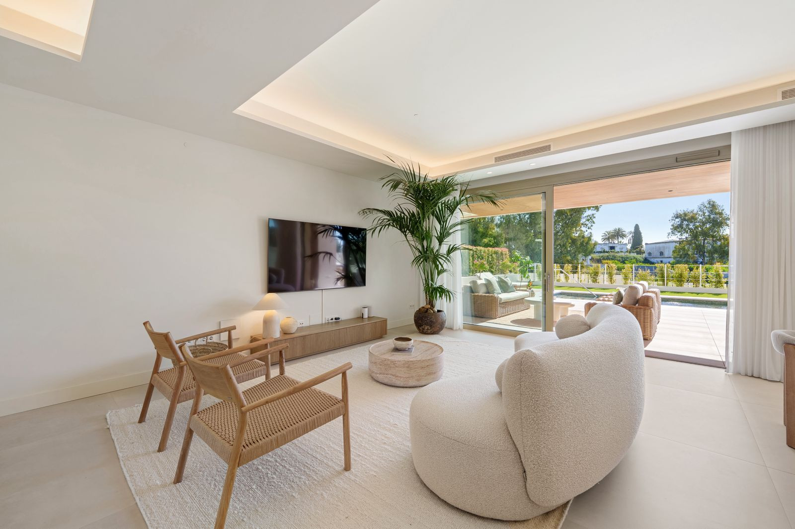 Modern living room with a large screen TV, white sofa, two wooden chairs, and a sliding glass door.