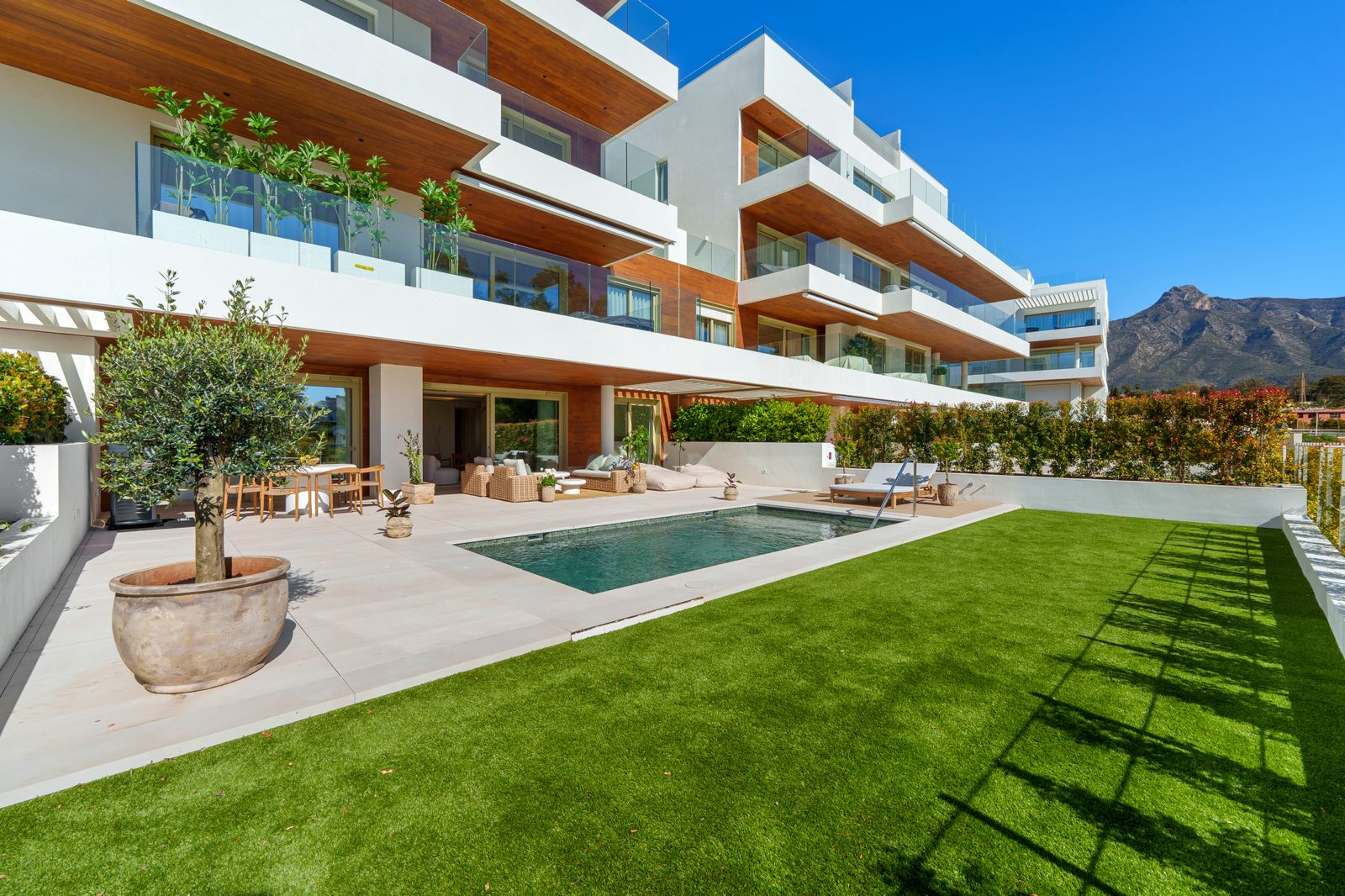 Contemporary building exterior with pool, artificial lawn, and mountains in the background under a blue sky.