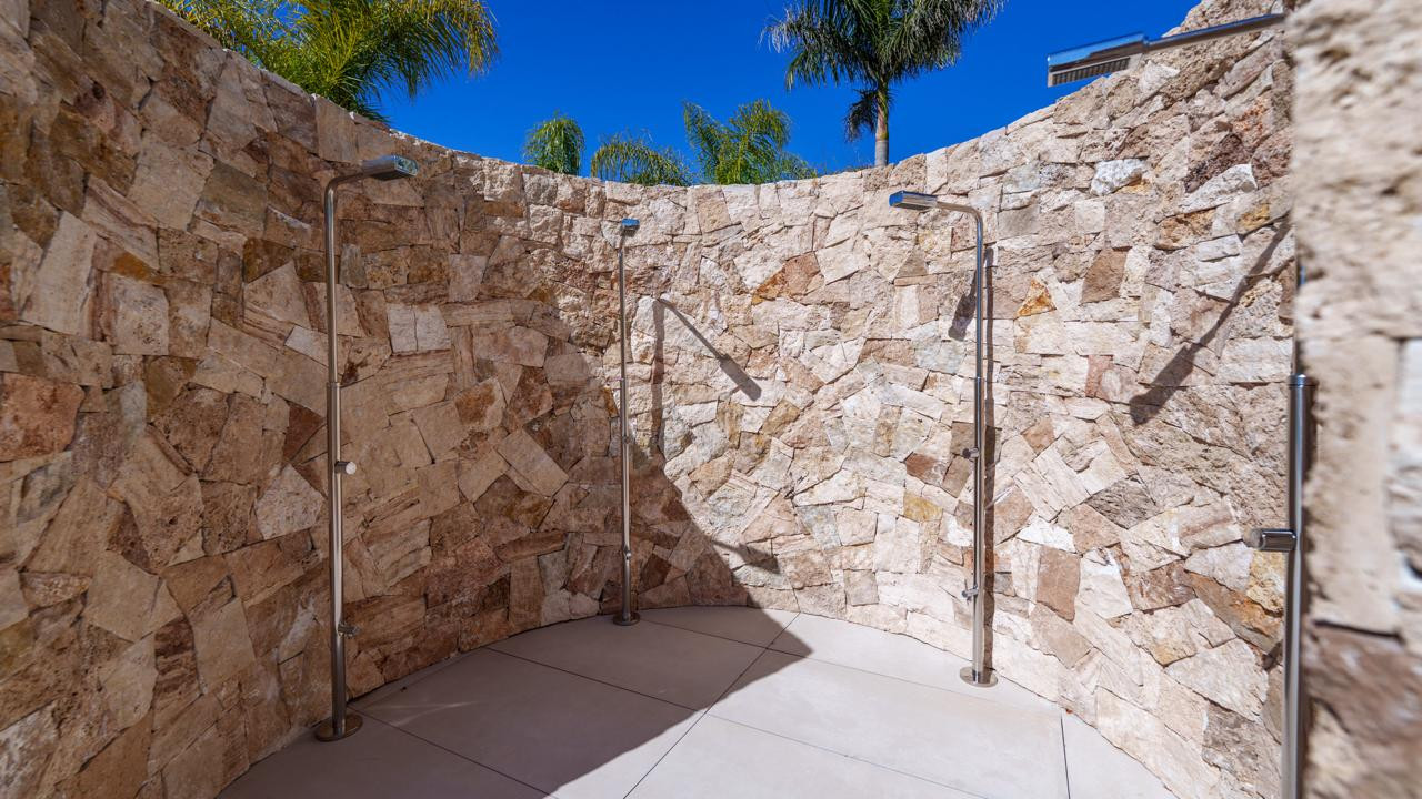 Curved outdoor shower area with stone walls, multiple shower heads, and a light-colored floor.