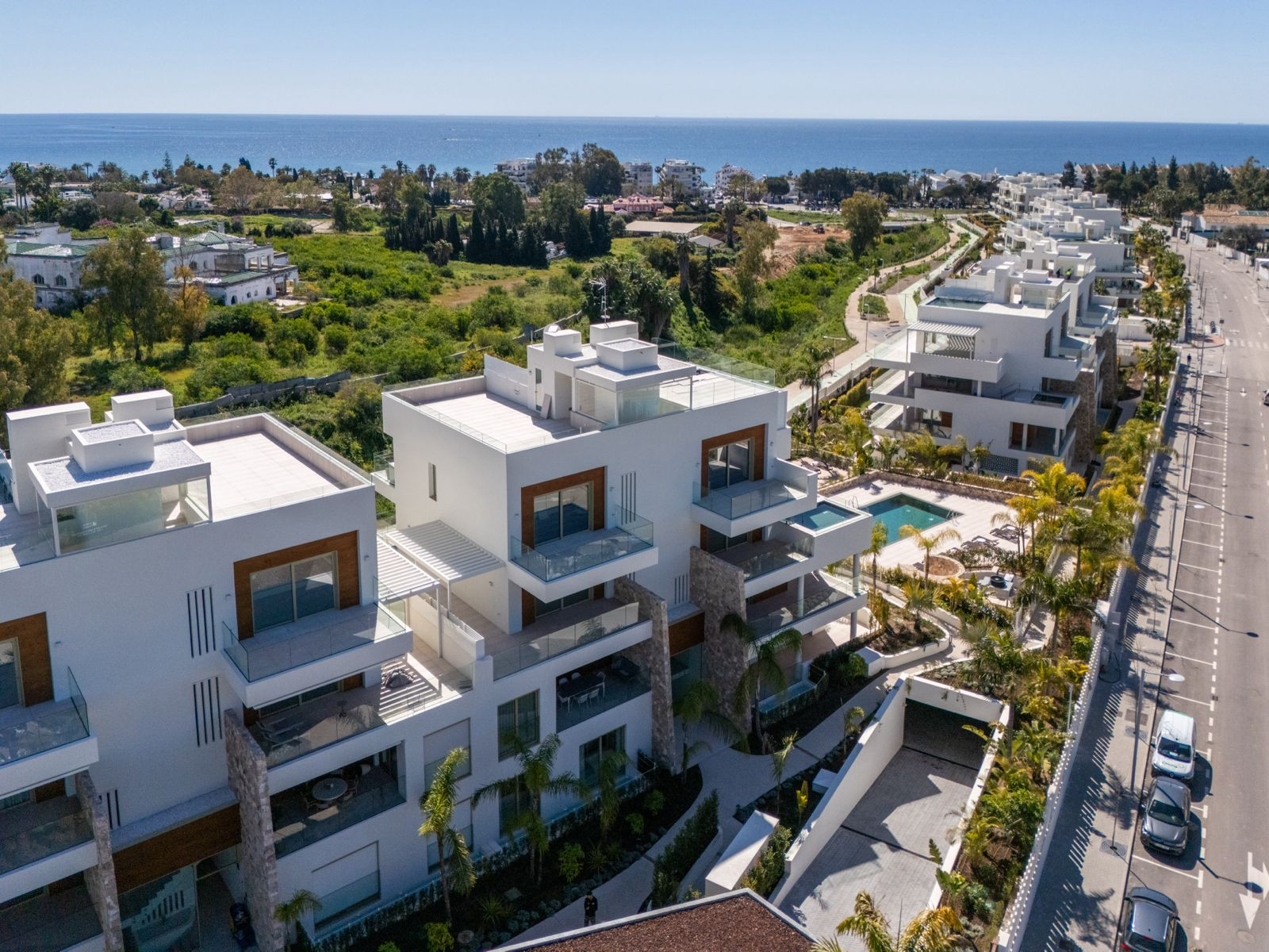 High aerial view of multiple white modern apartment buildings with balconies, a pool, and ocean in background.