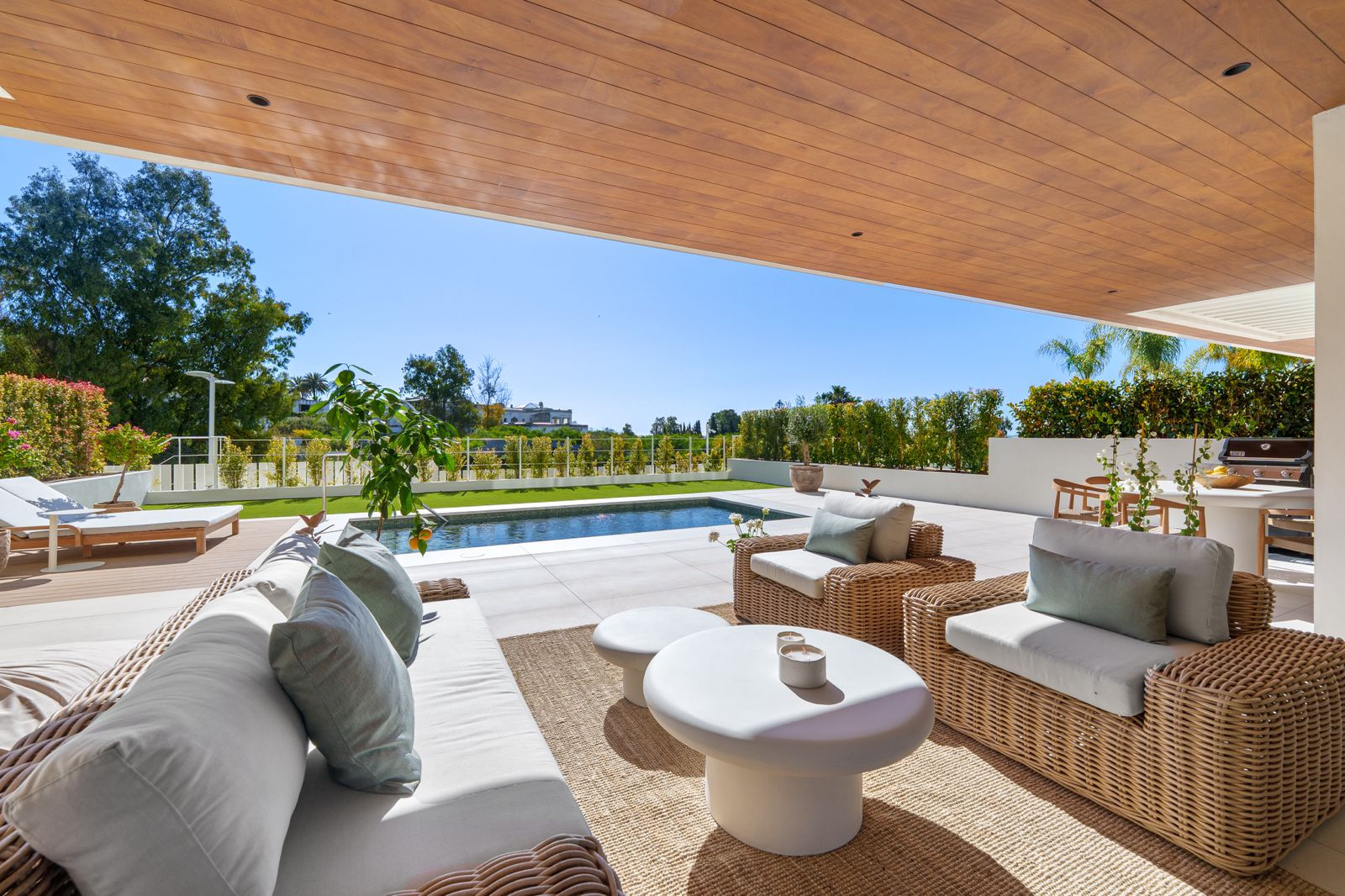 Covered outdoor patio with a seating area, overlooking a rectangular pool and a green lawn.