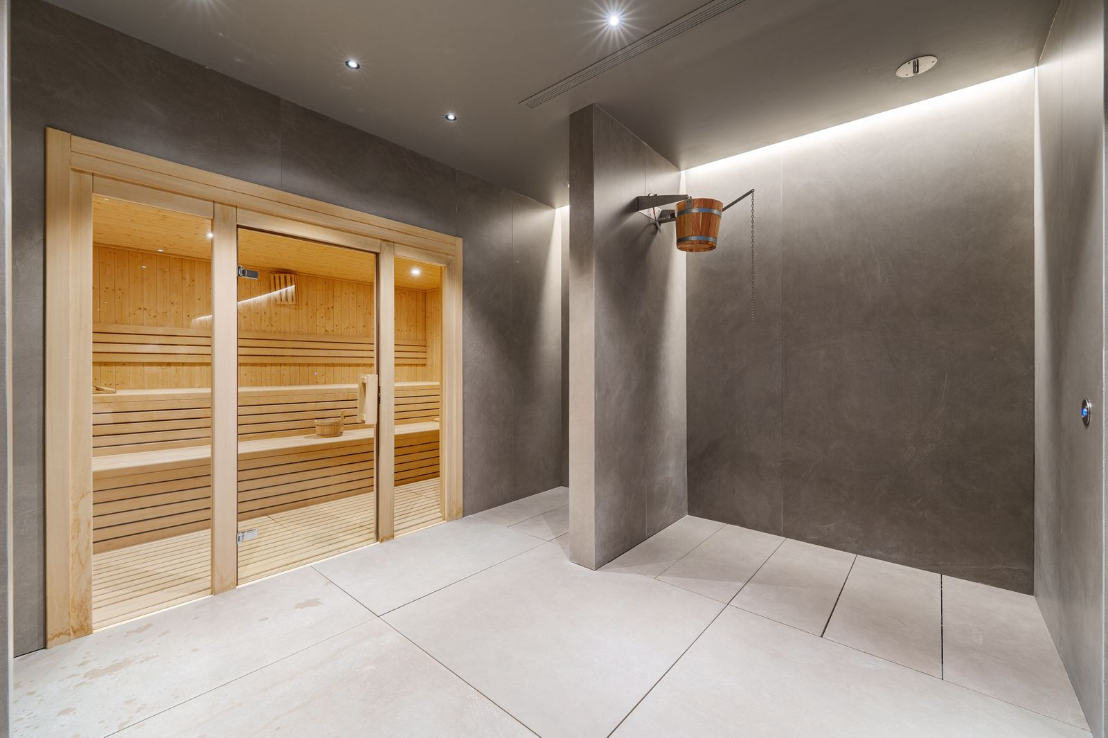 Spa area with a wooden sauna cabin, a cold plunge bucket shower, and grey tiled walls and floor.