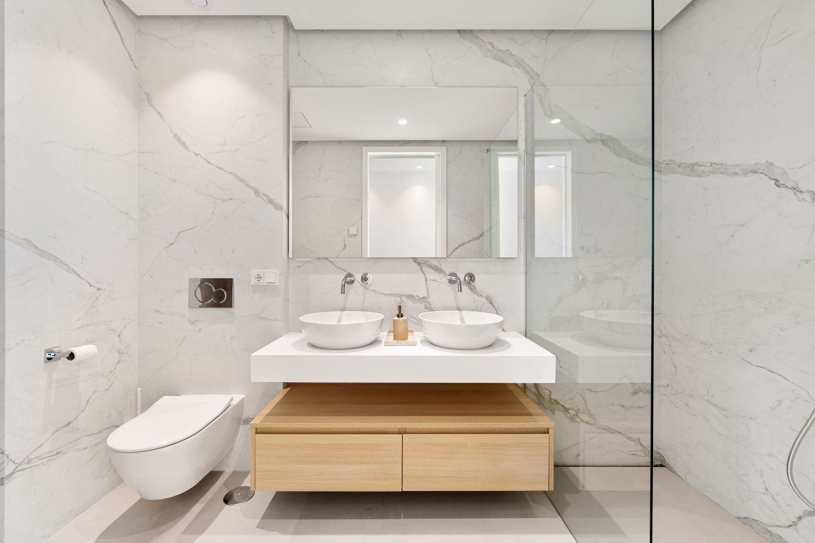 Bathroom with dual sinks, marble walls, a wall-mounted toilet, and a glass shower enclosure.