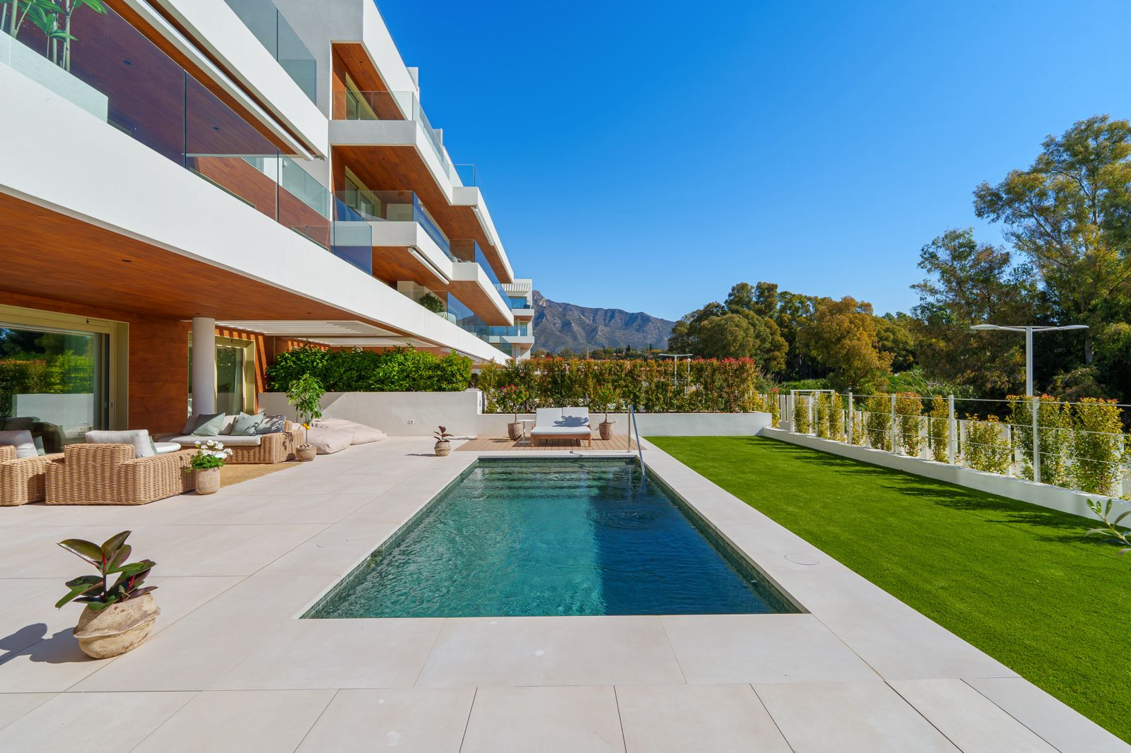 Outdoor pool area with grass lawn, modern building facade, mountains in the distance, and clear blue sky.