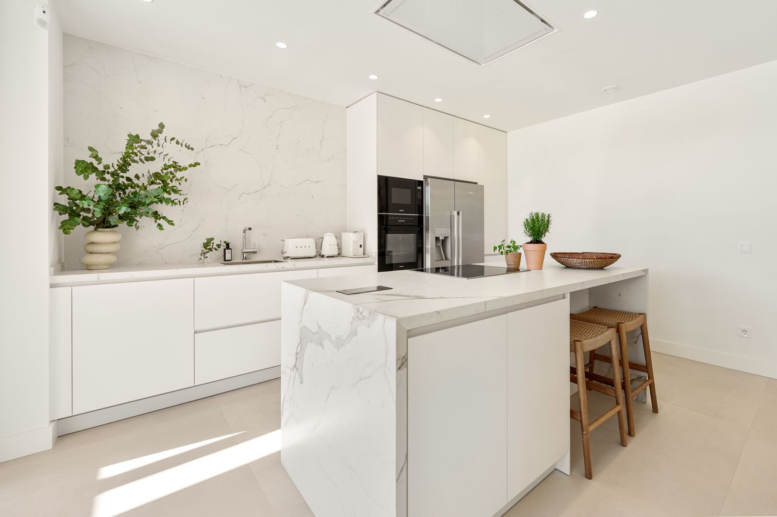 Modern white kitchen with a marble island, matching countertops, integrated appliances, and potted plants.