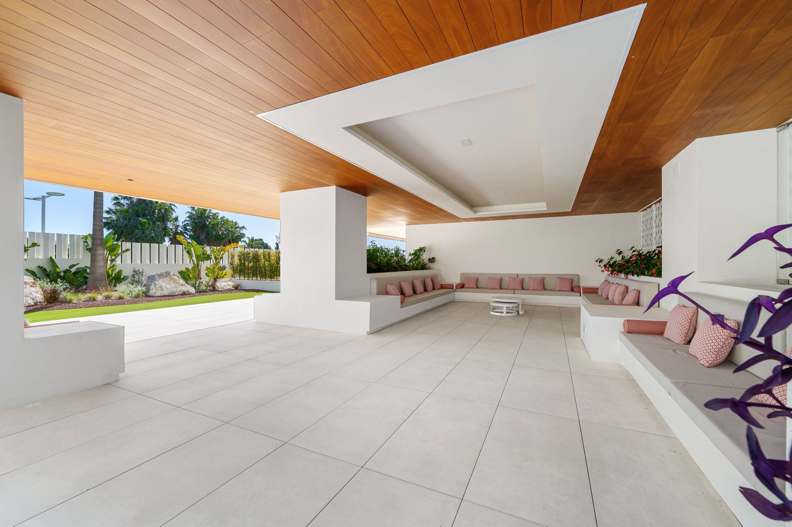 Covered outdoor lounge area with built-in seating, pink cushions, white tiled floor, and wooden ceiling.