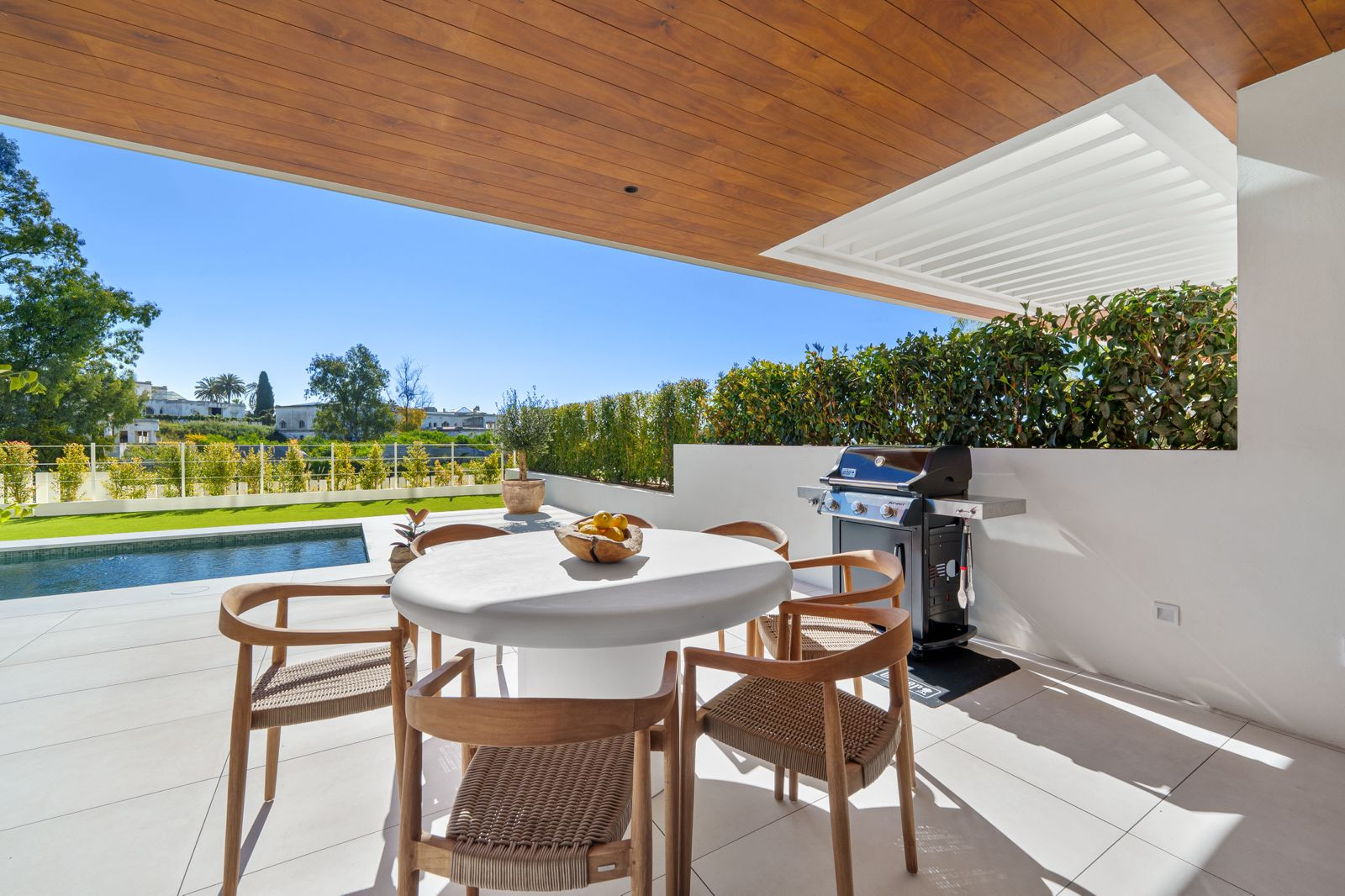 Covered outdoor dining area with a round white table, six chairs, and a grill, next to a pool.