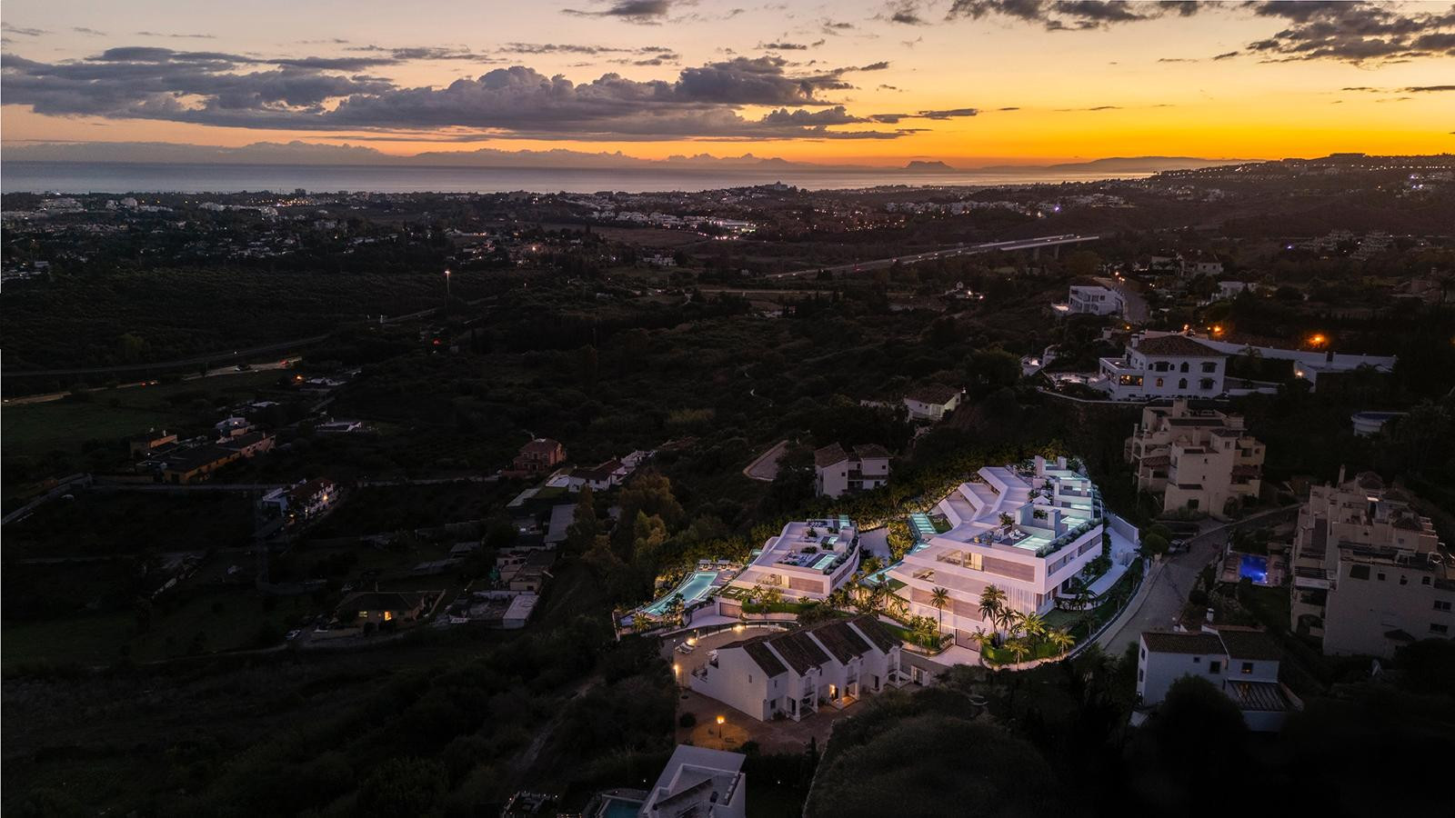 Aerial view of a modern apartment complex nestled into a hillside at dusk, with lights illuminating the buildings.