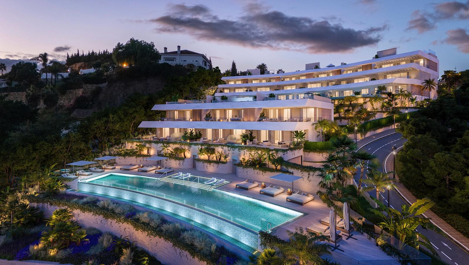 Exterior view of a multi-story apartment complex at dusk, with a large infinity pool and many lounge chairs.