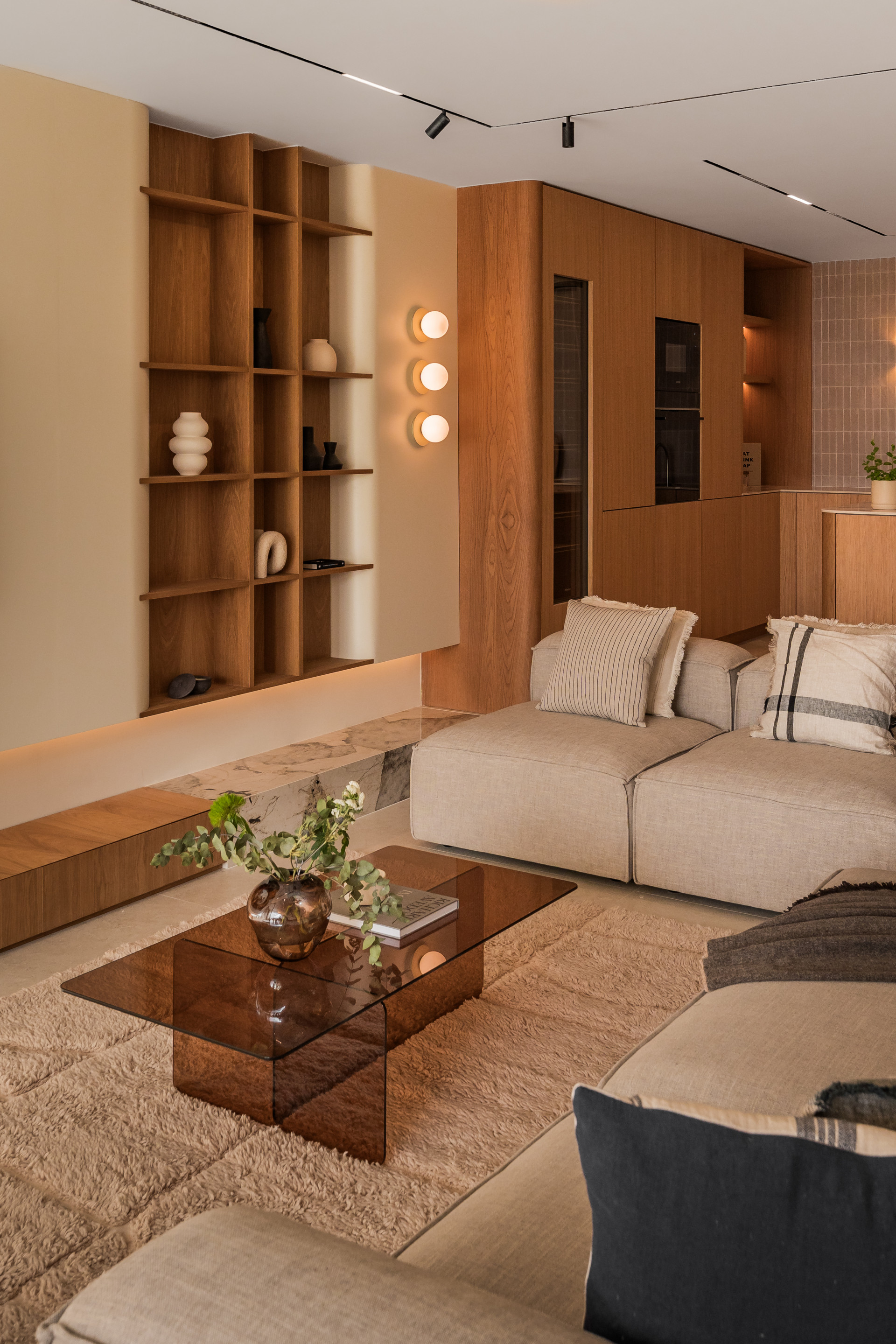 Living room with beige sectional sofa, glass coffee table, and built-in wooden shelving against a light wall.