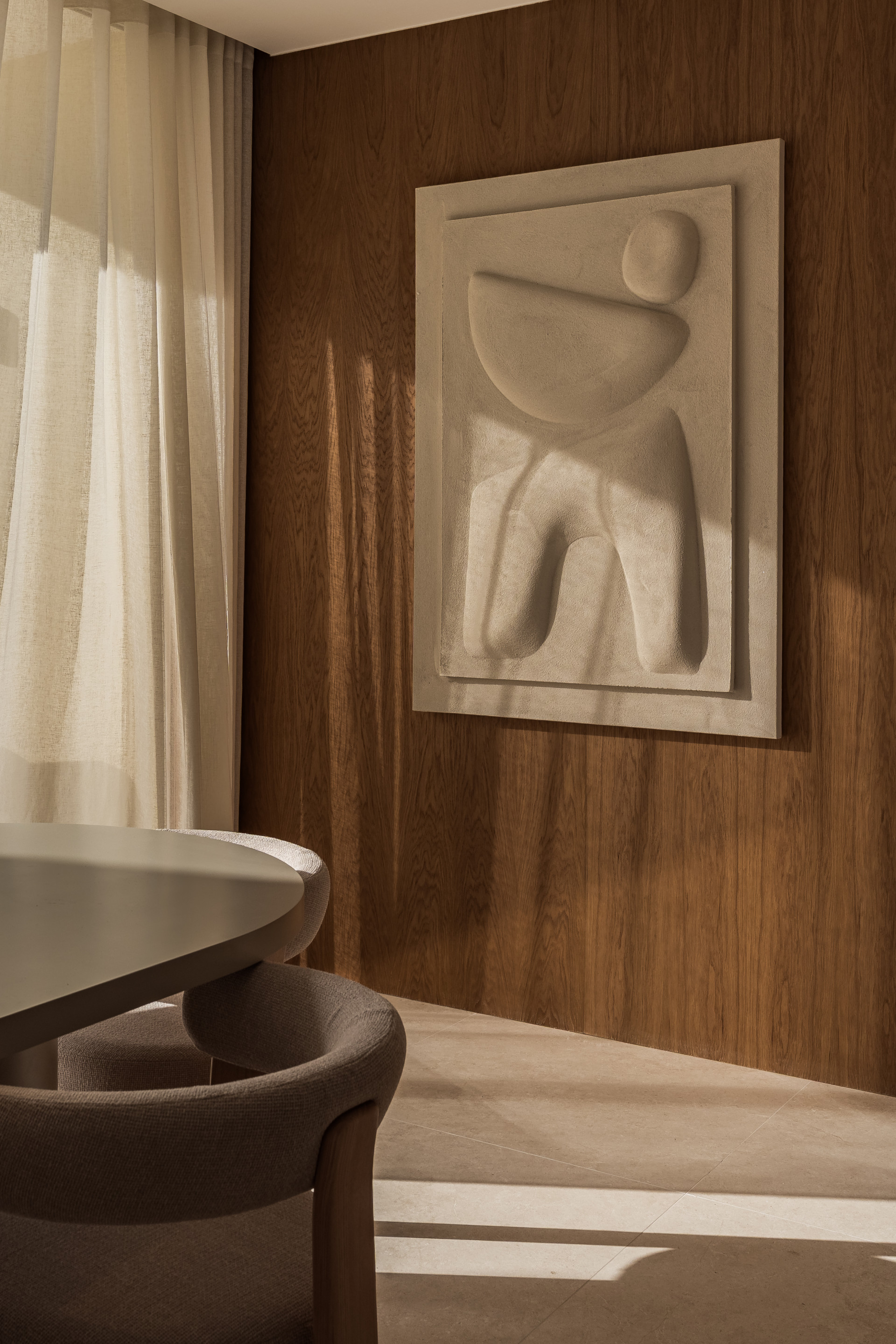 Dining area with a corner of a table and chairs, illuminated by sunlight filtering through sheer curtains.