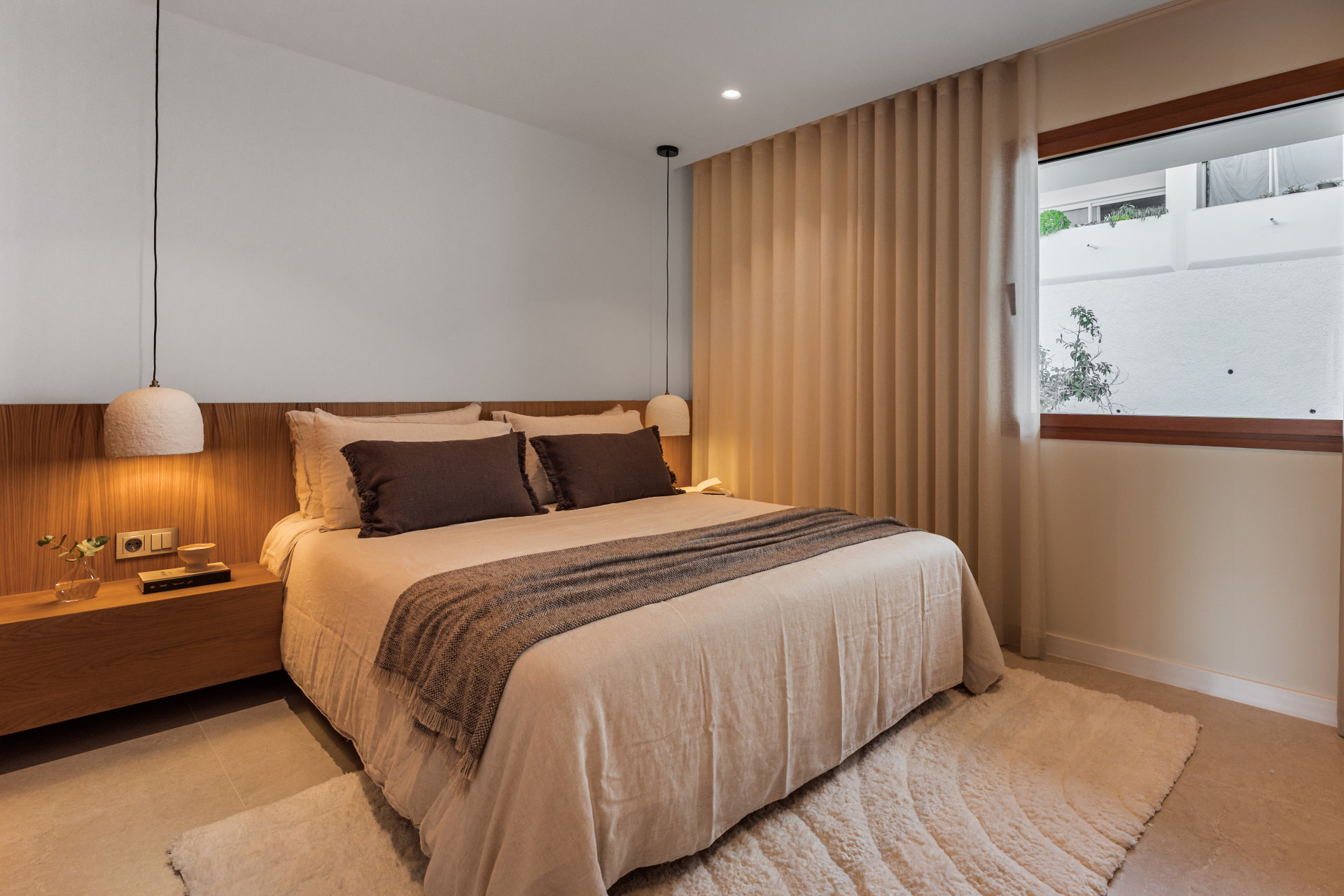 Bedroom with a large bed, two hanging pendant lights, a wooden headboard, and a rectangular window.