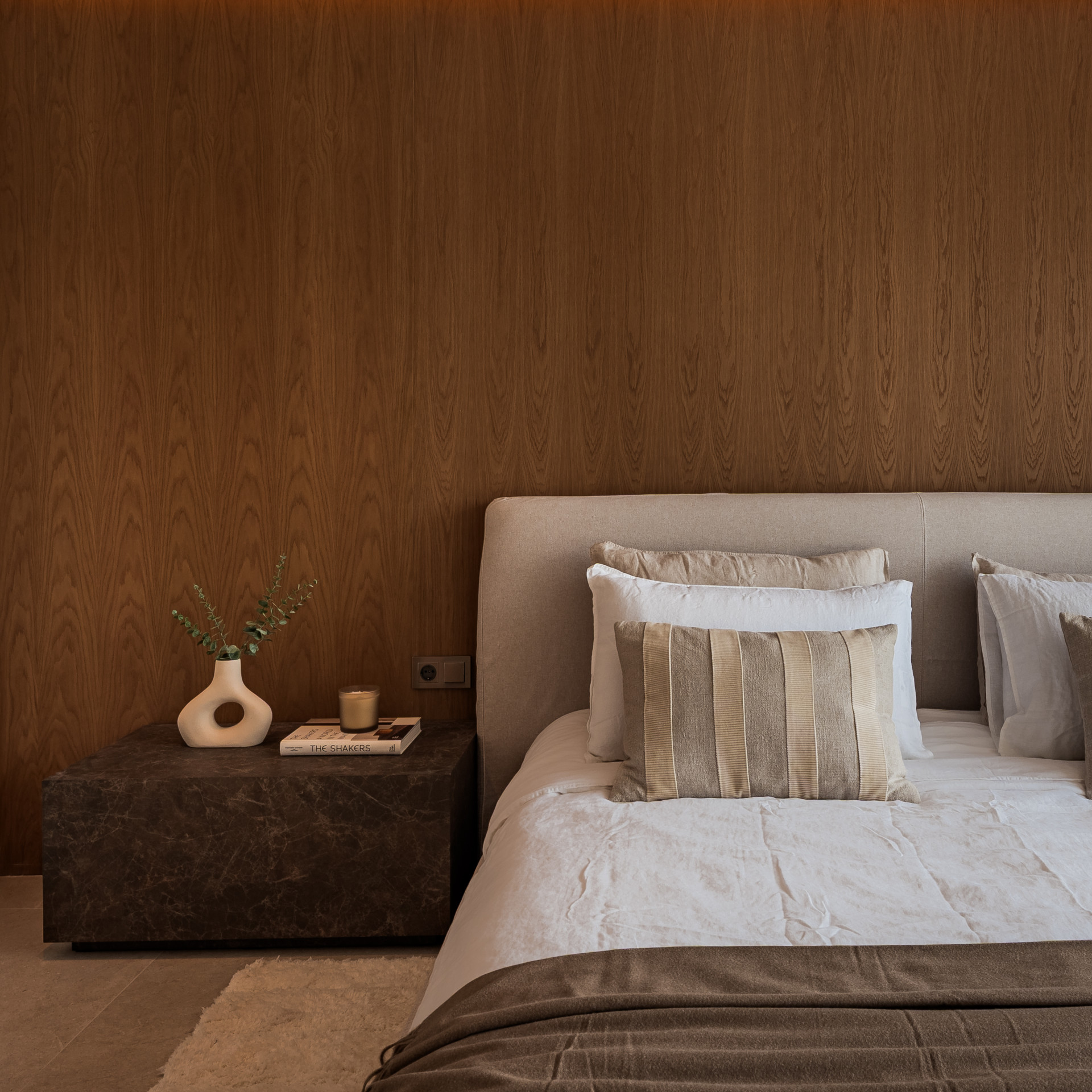 Close up of a bed with white and beige pillows, a dark brown nightstand, and a wooden paneled wall.