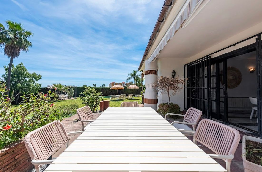 Outdoor patio with a large white dining table and chairs, overlooking a garden and distant pool area.