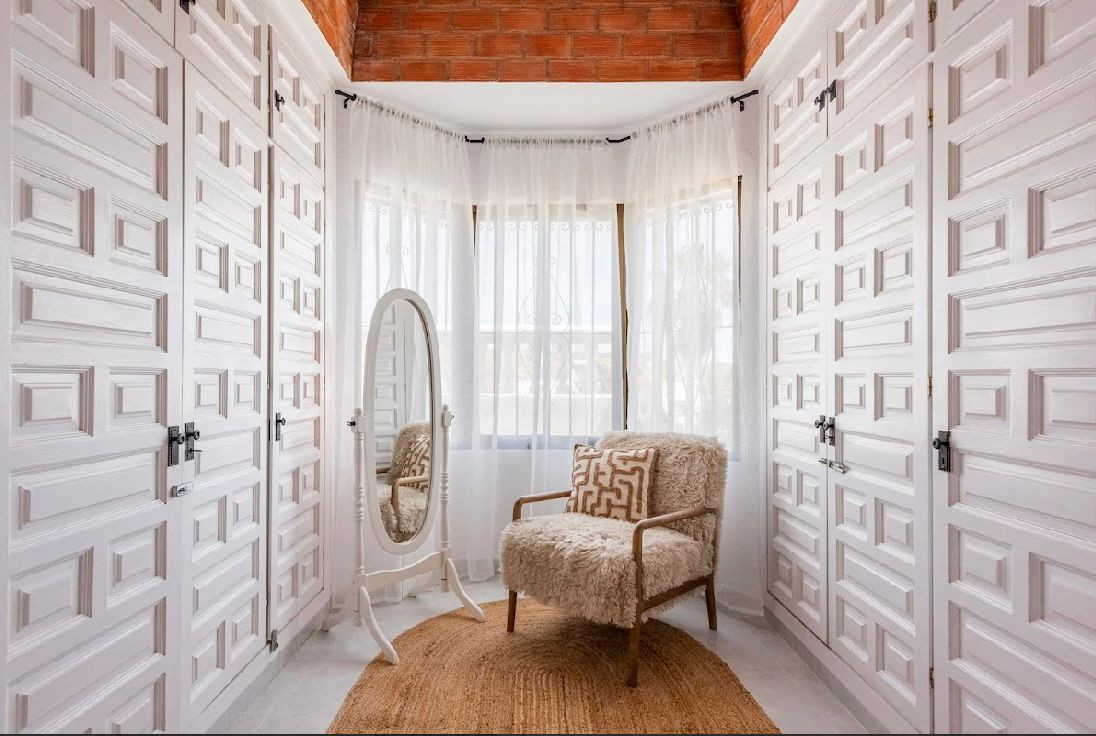 Walk-in closet with white paneled doors, full length mirror, shaggy armchair, and sheer curtains.