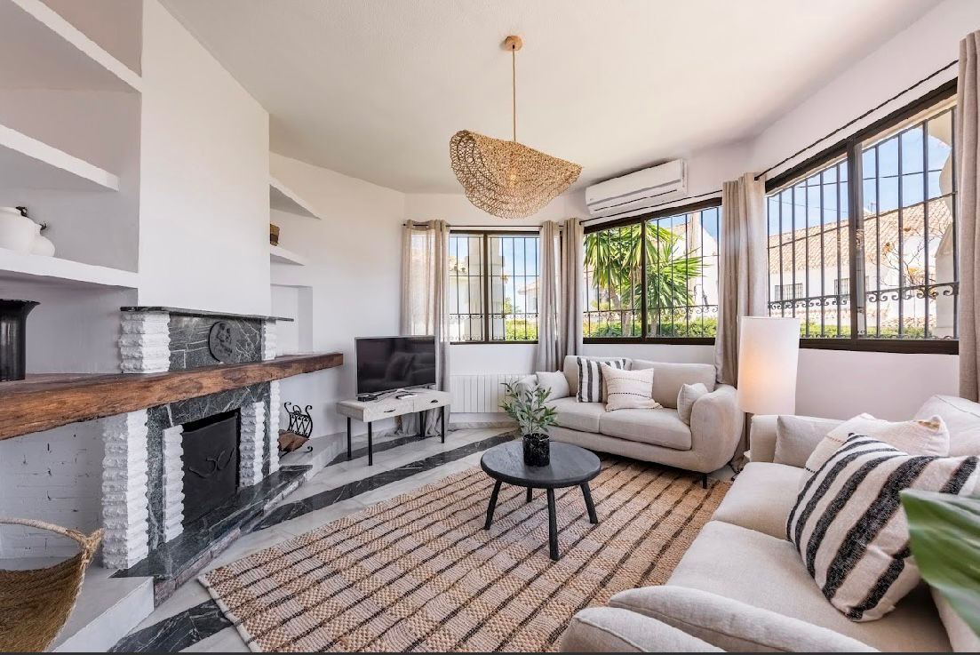 Living room with a fireplace, two light sofas, a textured rug, and large windows with iron grilles.