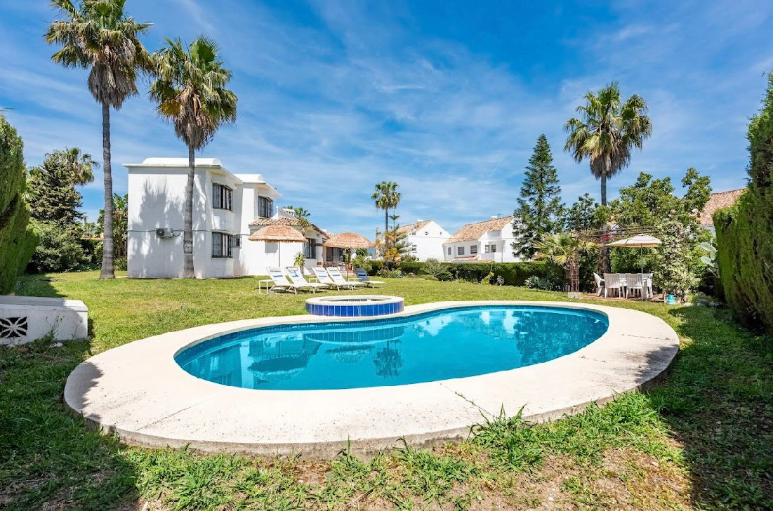 Oval swimming pool in a green lawn, with a white villa, palm trees, and straw umbrellas in the background.