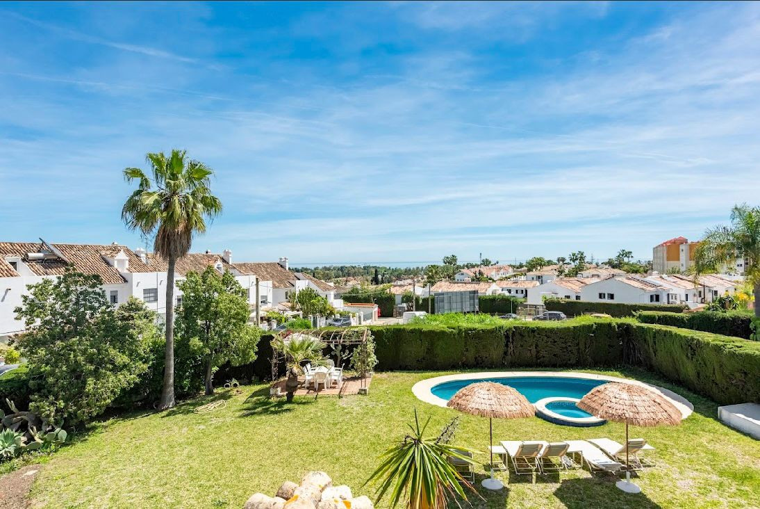 Aerial view of a villa's backyard with a pool, lounge chairs, outdoor dining, and distant town and sea.