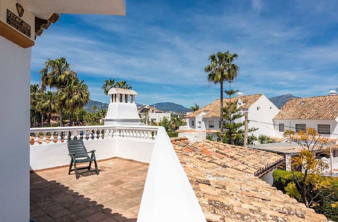 Rooftop terrace overlooking tiled roofs, palm trees, mountains, and other houses under a blue sky.