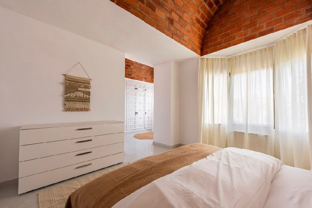 Bedroom with a white dresser, arched brick ceiling, large windows with sheer curtains, and a door.