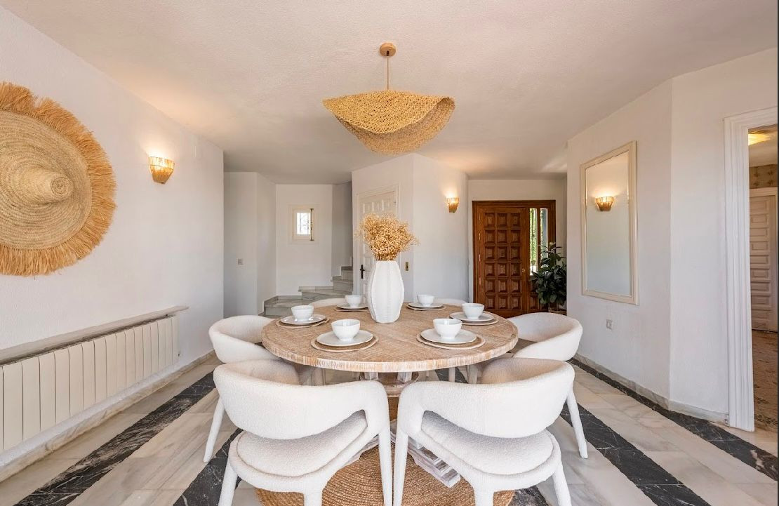 Dining room with a round wooden table, six white chairs, striped marble floor, and straw wall decor.