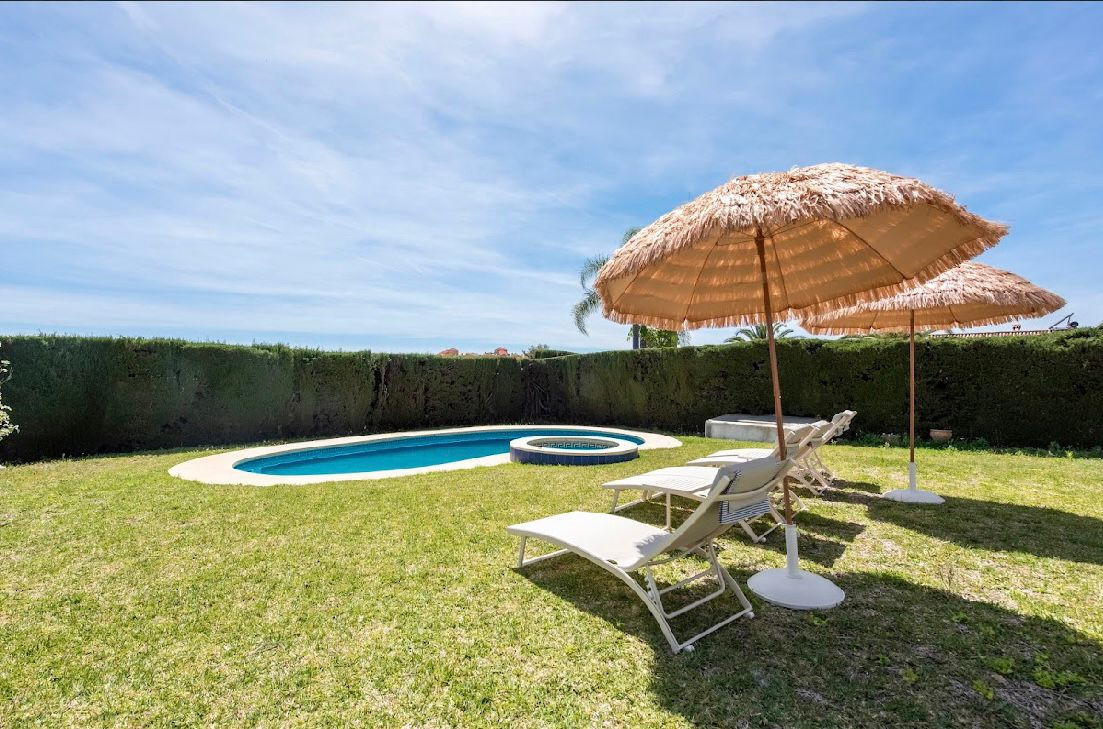 Outdoor view with a freeform swimming pool, two straw umbrellas, and several sun loungers on a green lawn.