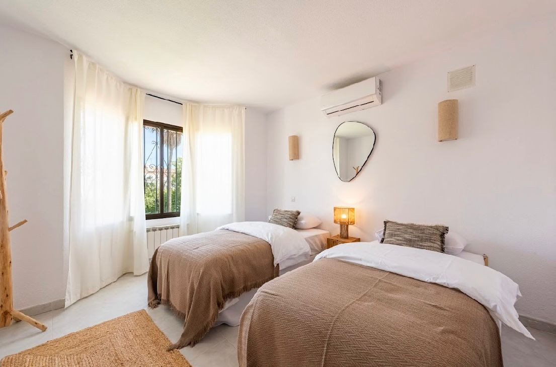 Bedroom with two single beds, air conditioning unit, a wall mirror, and two windows with curtains.