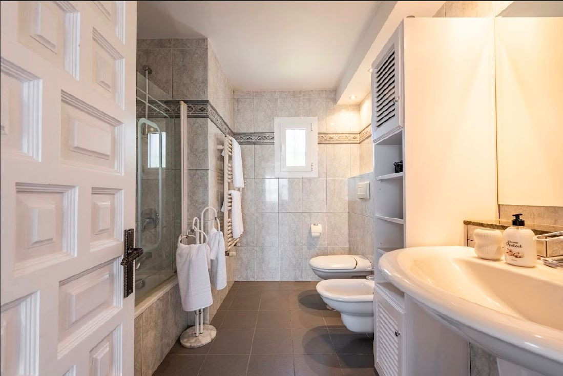 Bathroom with tiled walls, shower over tub, two toilets, and a white vanity with sink.