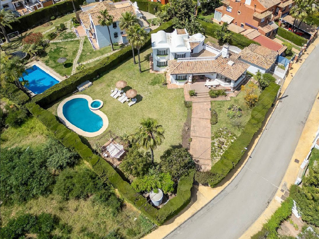 Aerial view of a white villa with two swimming pools, surrounded by green hedges and palm trees.