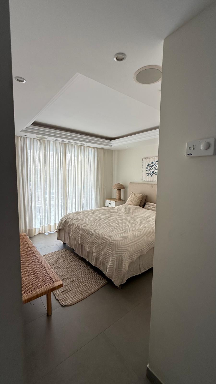 Spacious bedroom with a queen bed, woven bench, and sheer curtains allowing natural light.